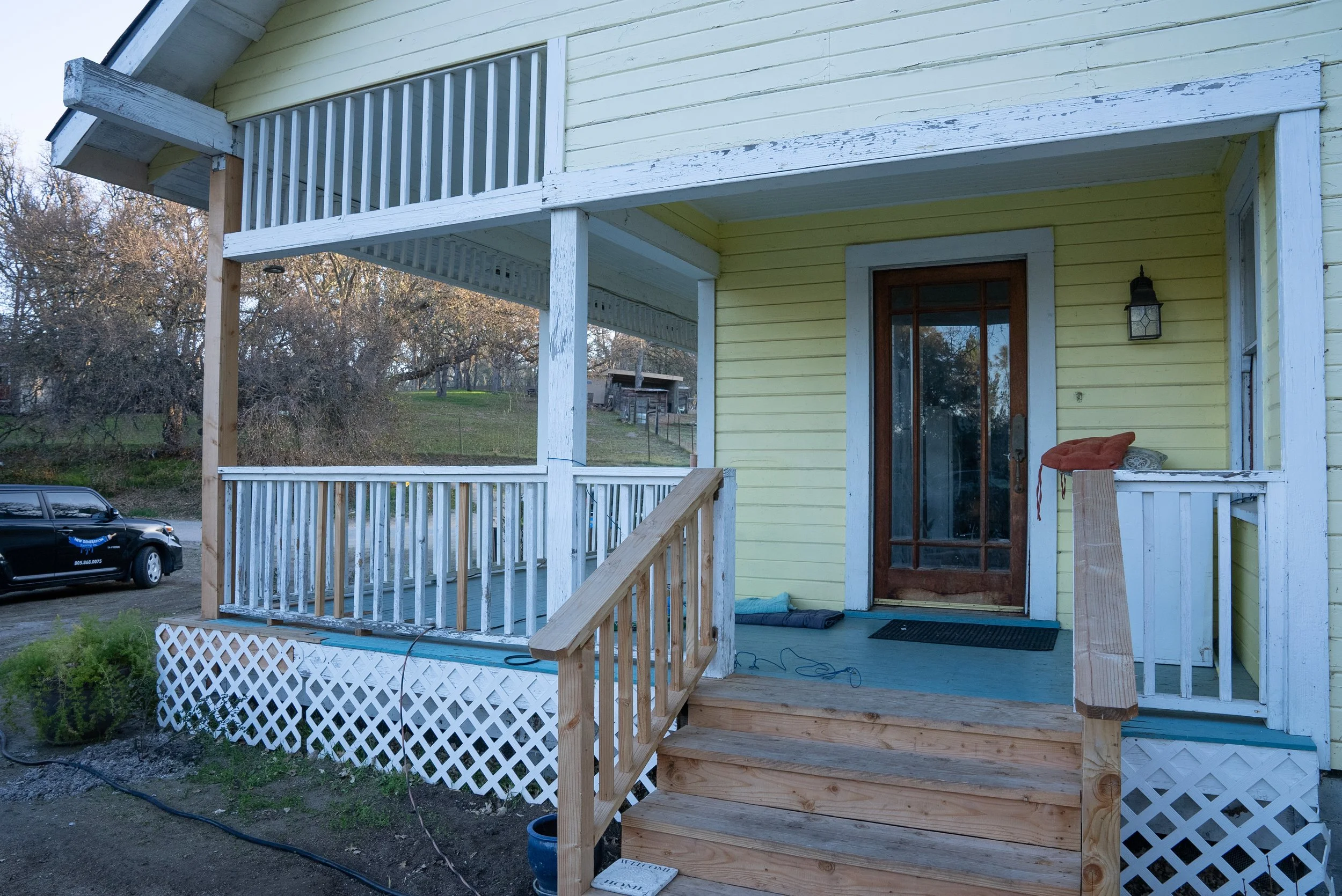 Front porch of a yellow house with white trim and a wooden staircase with railings, leading to a door with a glass panel. A black lantern-style light fixture is beside the door. There is a car parked nearby and some trees in the background.
