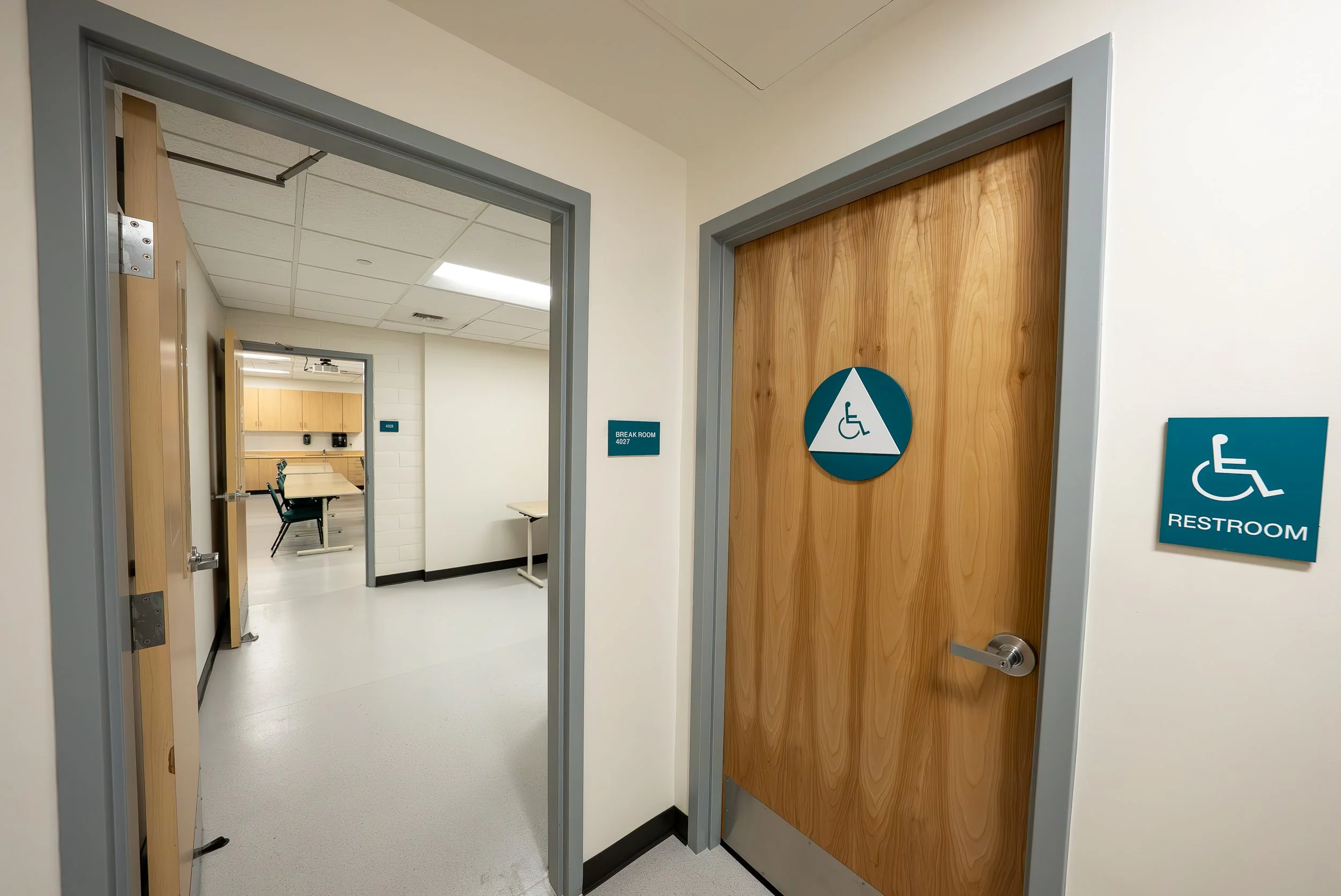 Hallway in a building with a door marked for accessible restroom, featuring a wheelchair symbol on the door and a sign indicating 'Break Room 4027'. There are tables and chairs visible through an open door in the background.