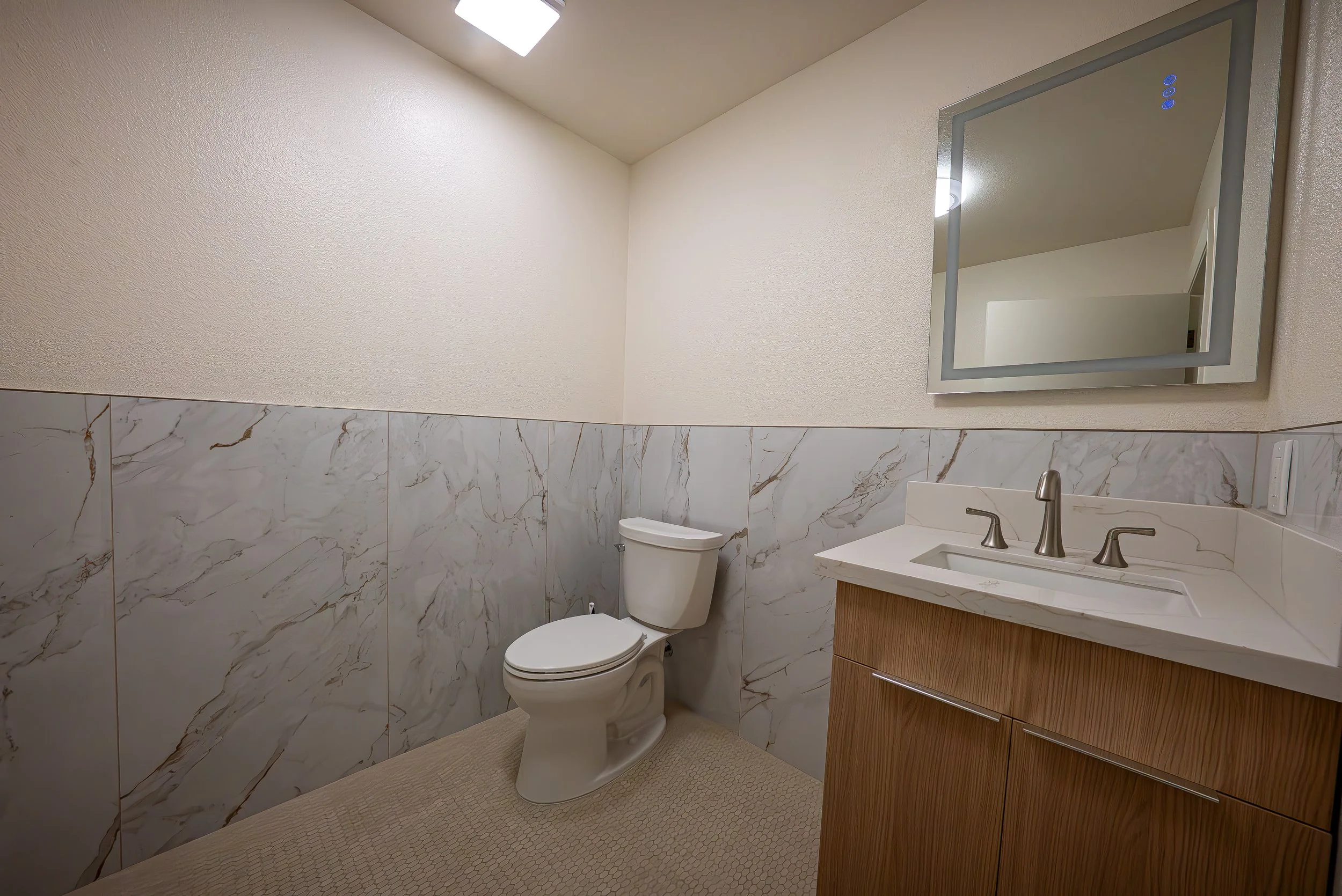 A bathroom with a toilet, sink with a wooden cabinet, mirror, marble tiled walls, a ceiling light, and beige textured flooring.