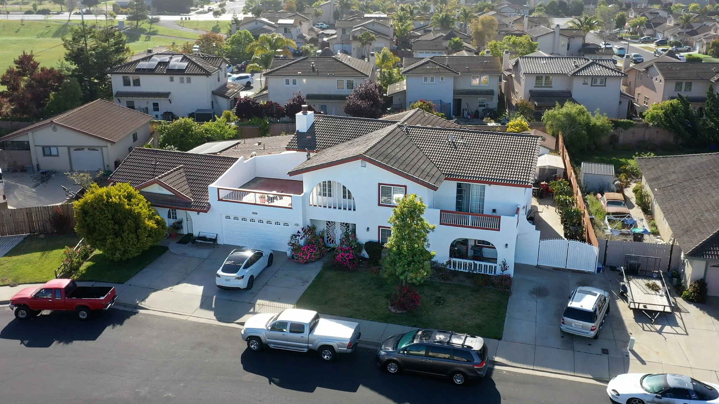 An aerial view of a residential neighborhood with multiple houses, including a prominent white house with a garage and a small front yard with pink flowers. There are cars parked in driveways and on the street, trees lining the properties, and neighb
