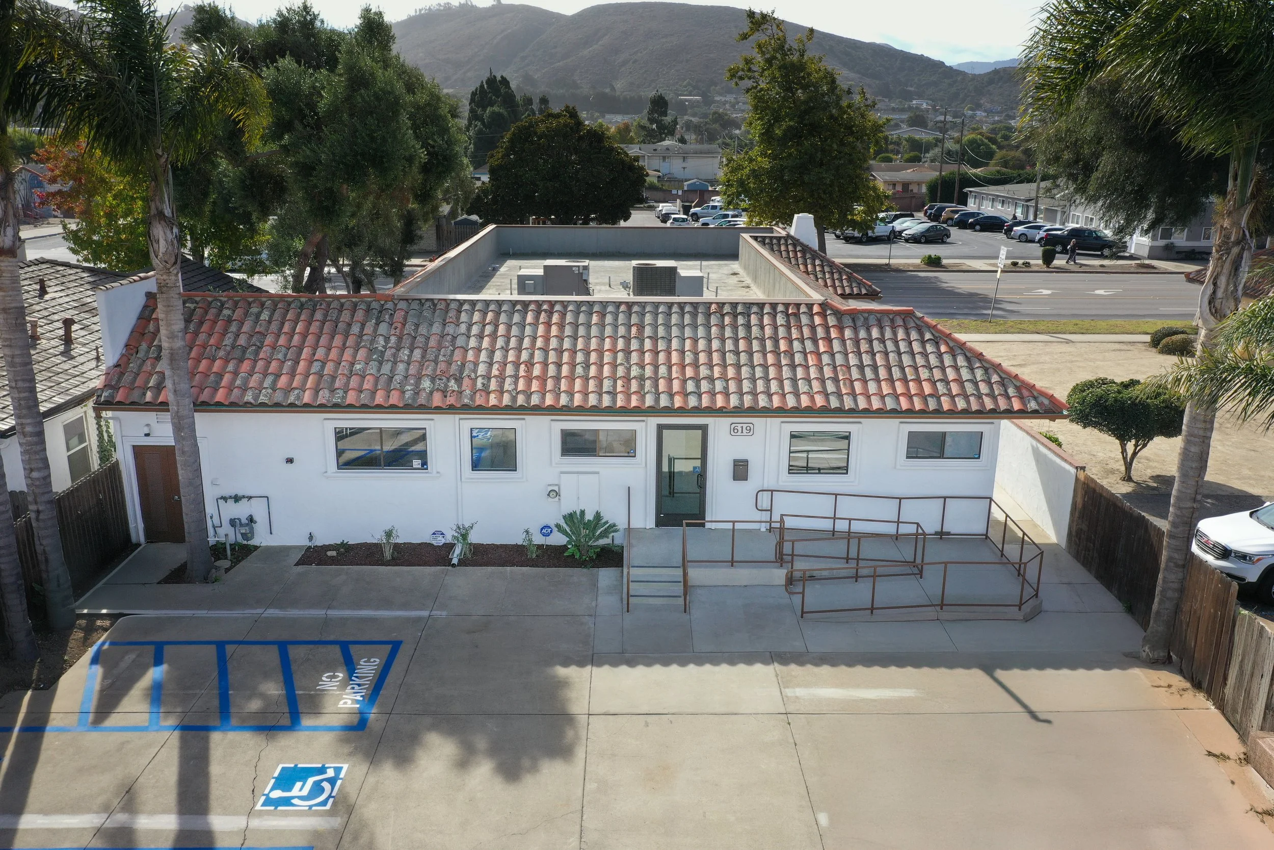 View of a small white building with a red clay tile roof, surrounded by trees and a parking lot with accessible parking spaces in the foreground.