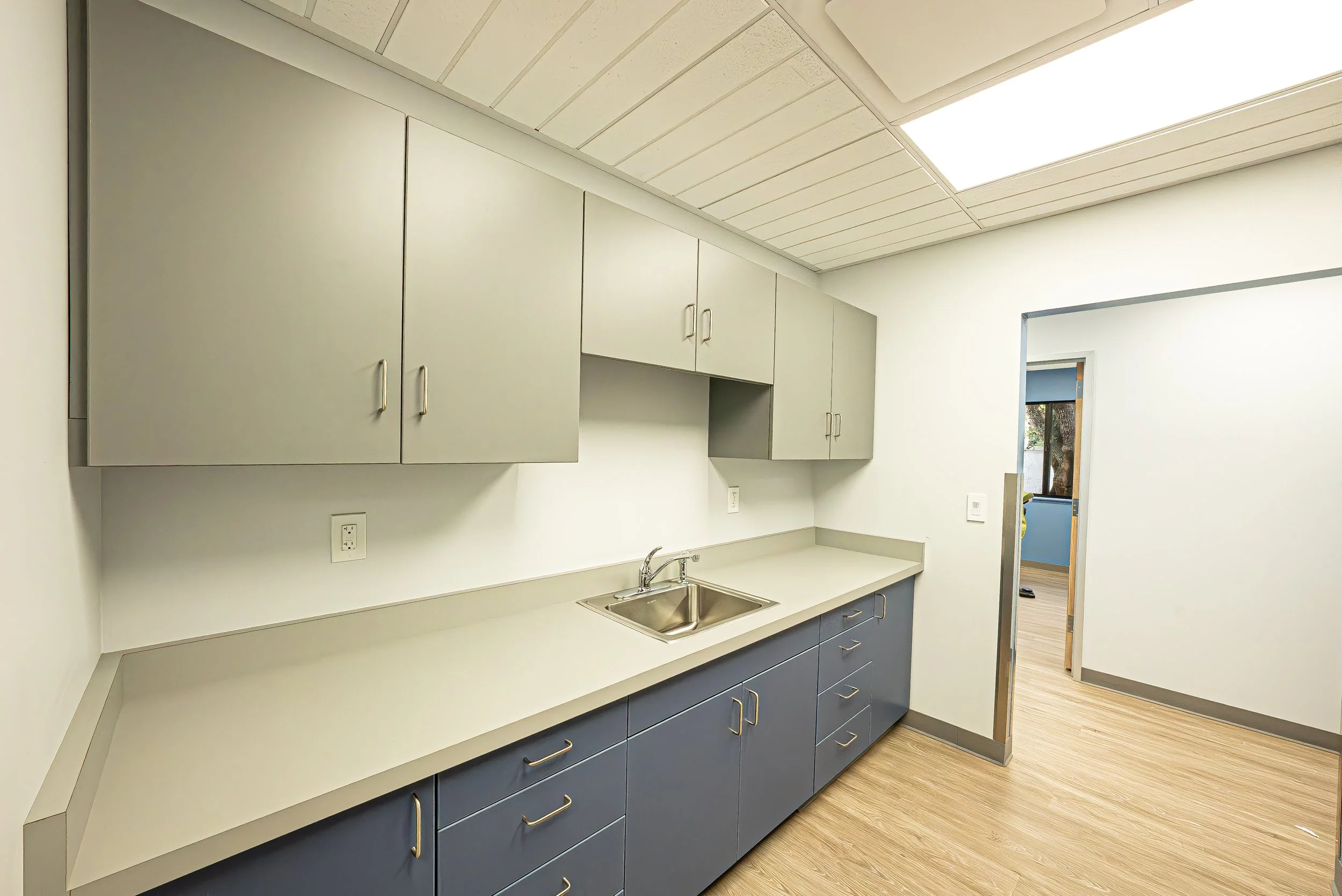A modern kitchen with blue cabinets, white countertops, a stainless steel sink, and upper cabinets. The room has a wood-like floor and a doorway leading to another room with a window.