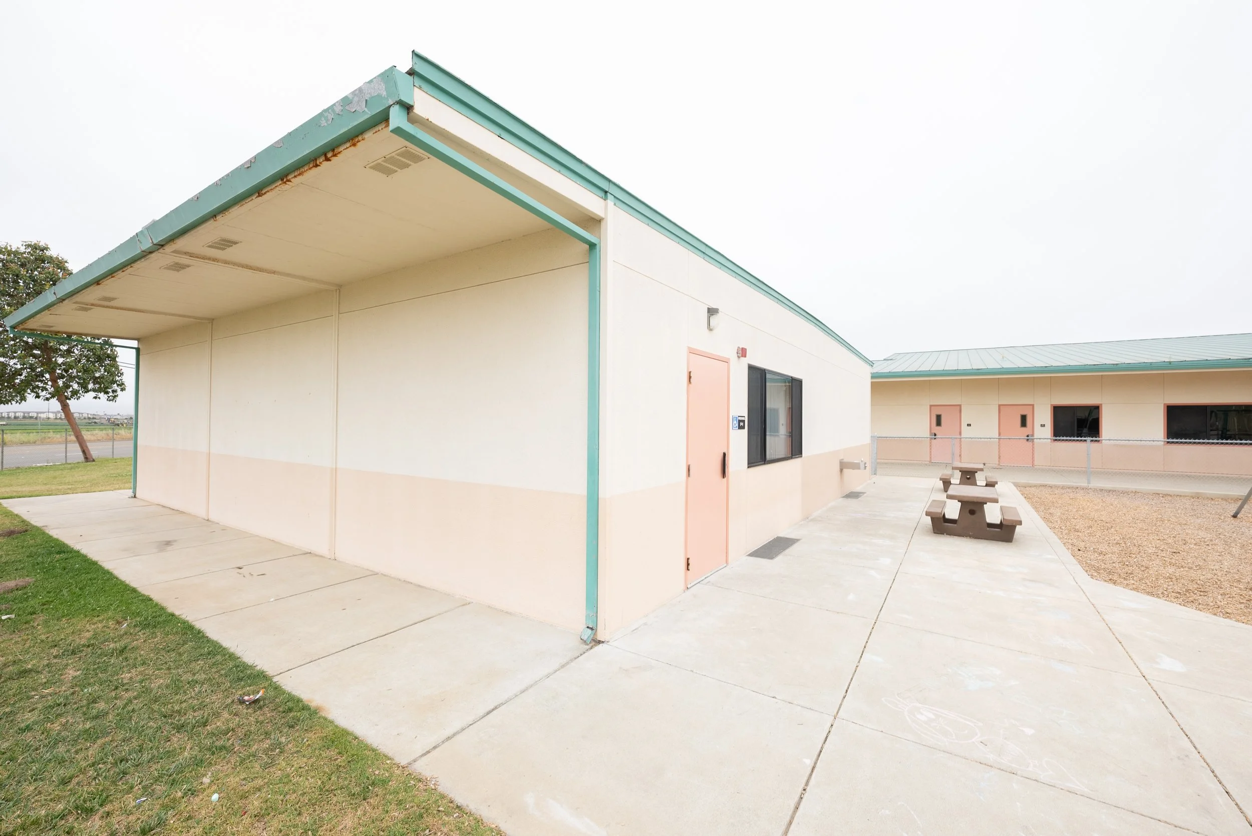An empty outdoor corner of a school or community center building with a small grassy area and a picnic table, under an overcast sky.