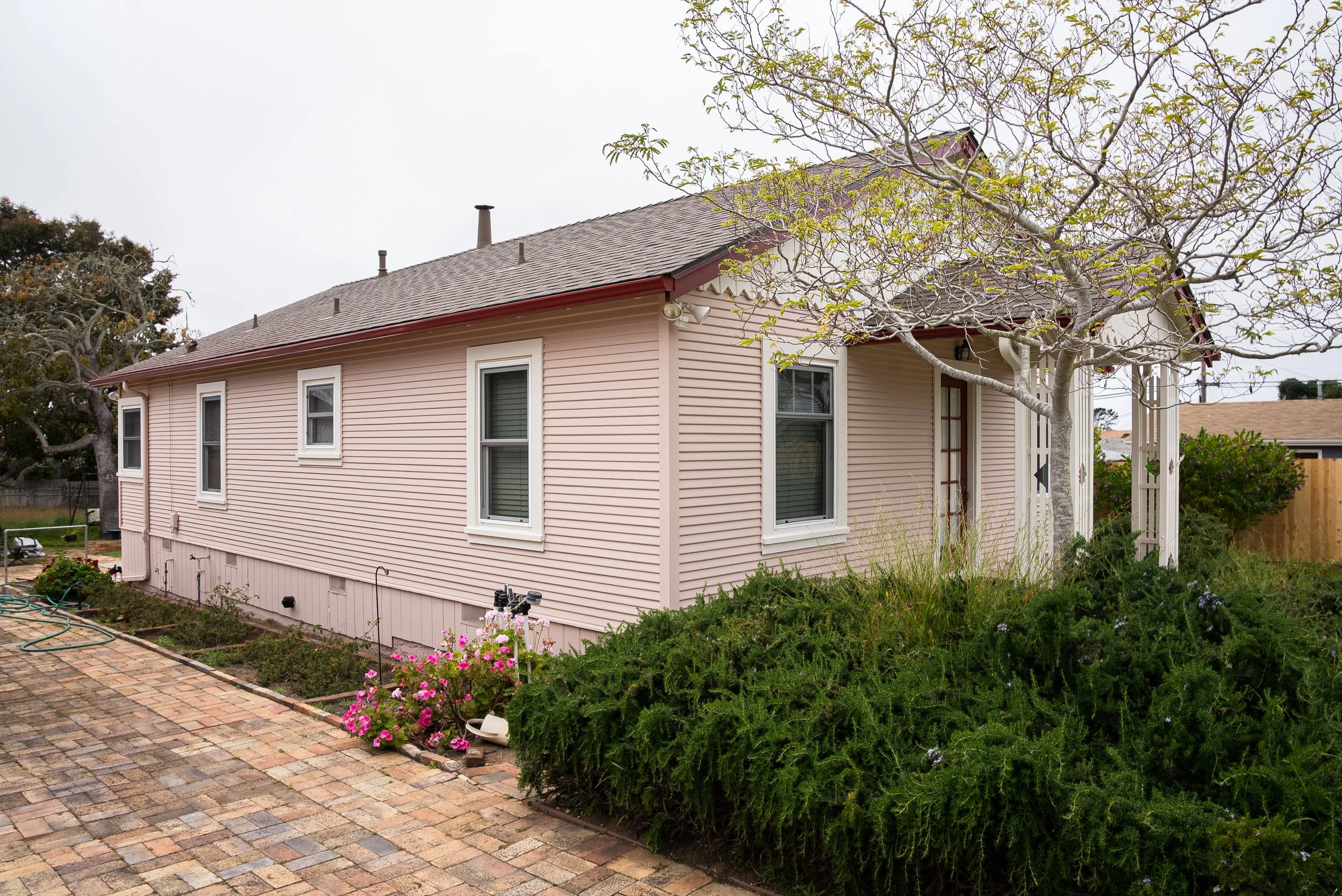 The side view of a pink house with a brick patio, some flowering plants, green shrubs, and leafless trees, with neighboring houses visible in the background.