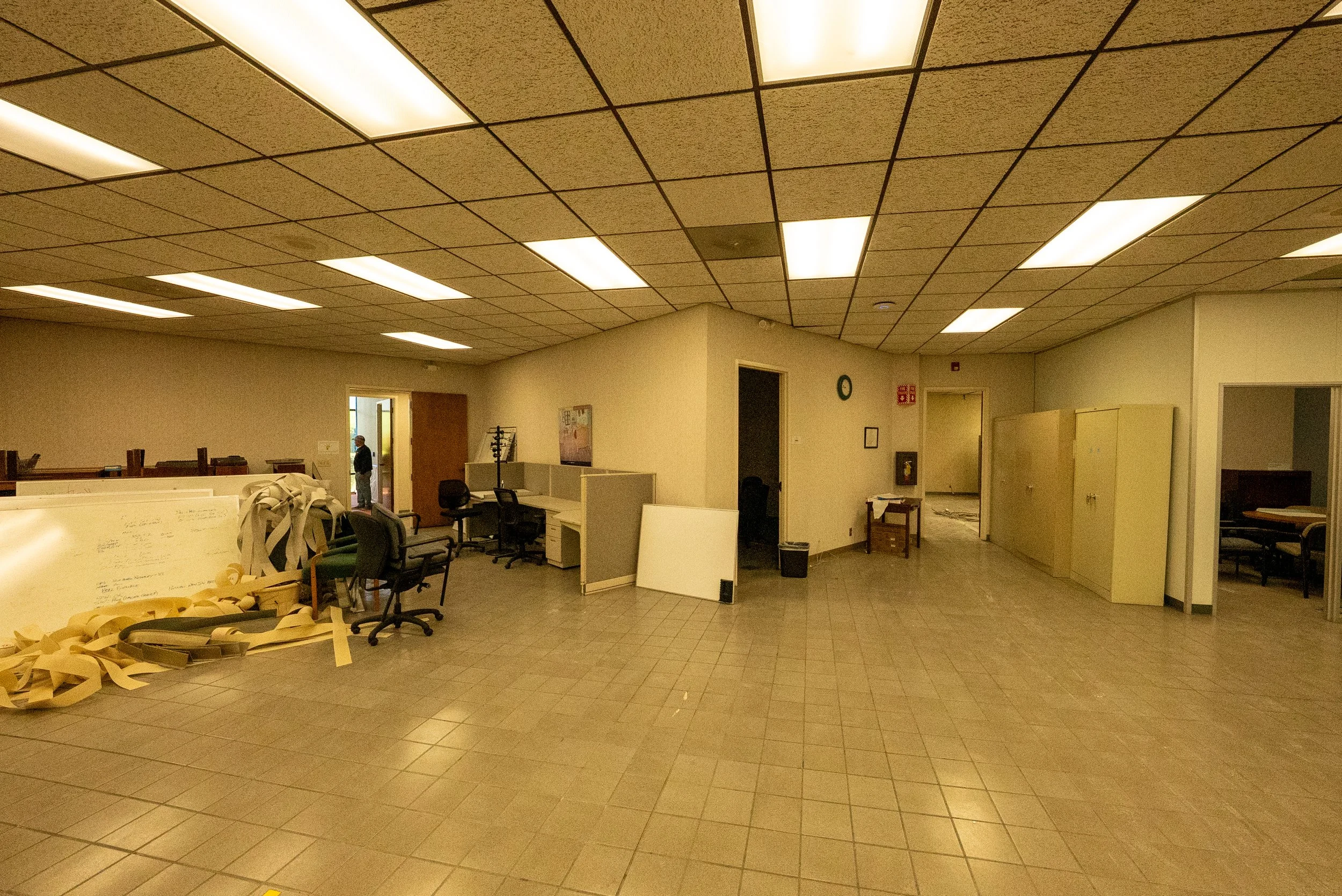 An office space with a suspended ceiling, fluorescent lights, cubicles, chairs, and some scattered office supplies and paper on the floor.