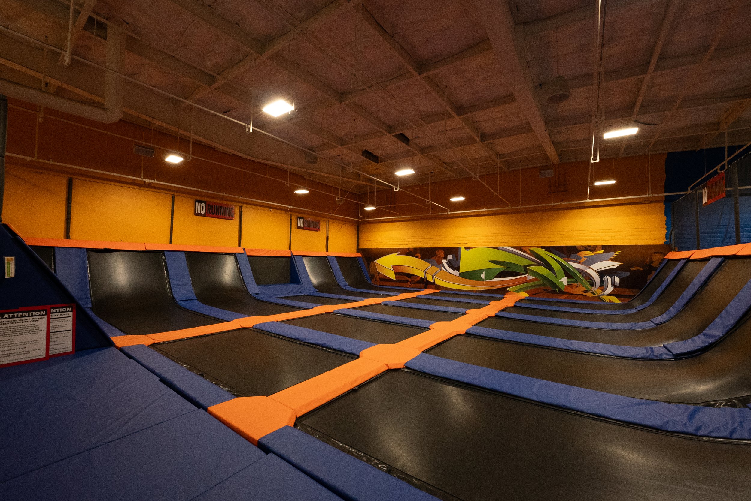 Indoor trampoline park with multiple trampolines lined up on a padded floor, graffiti art on the wall, and a sign that reads 'No Running'.