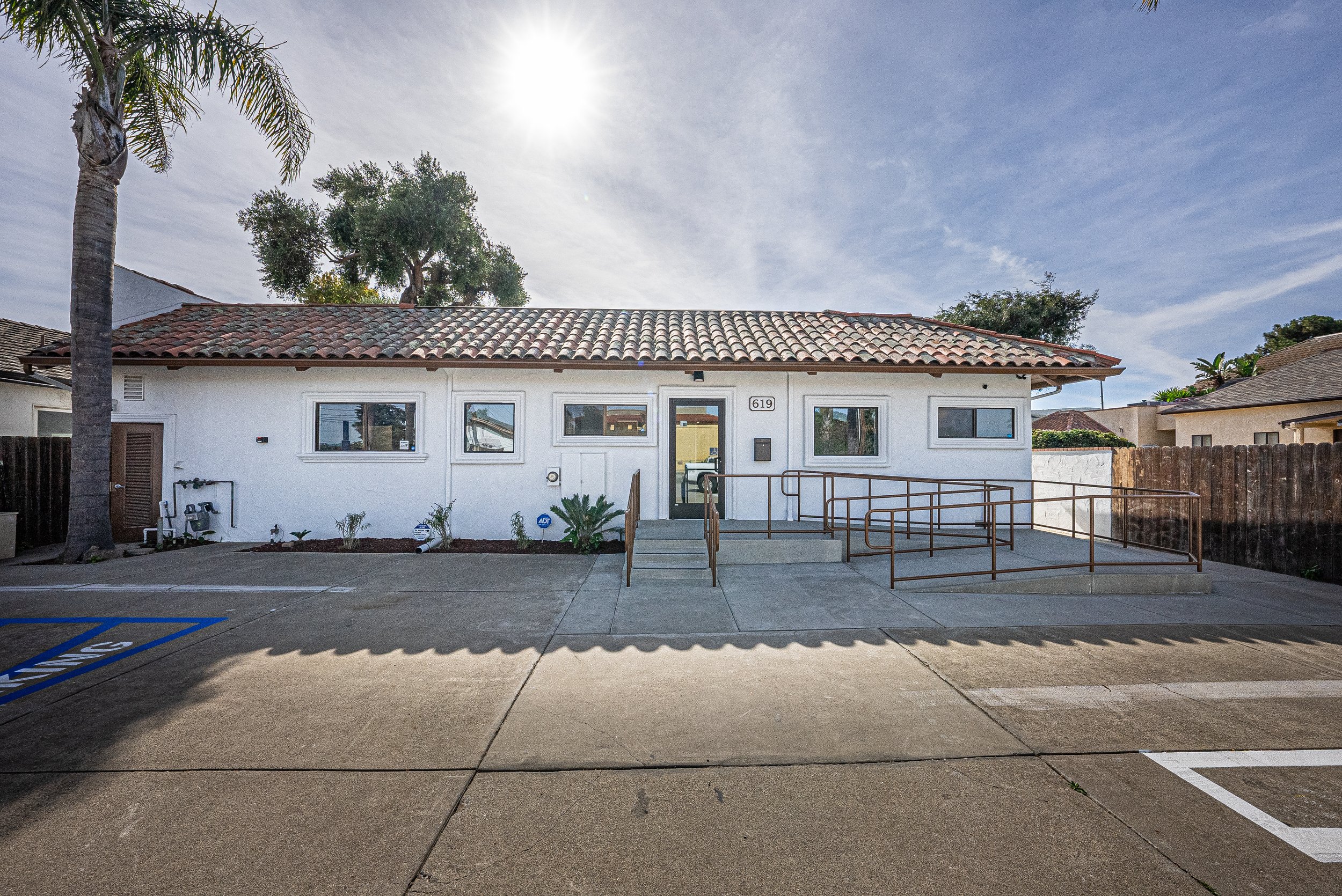 Front view of a single-story white building with a tiled roof, with palm and other trees in the background, and a parking lot in the foreground.