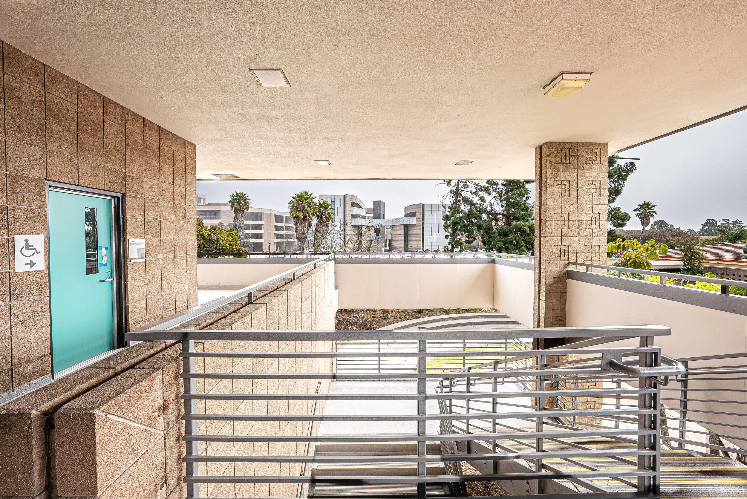 An outdoor balcony or walkway area with beige concrete walls and ceiling, metal railings, outdoor lighting fixtures, and a view of trees and modern buildings in the background.