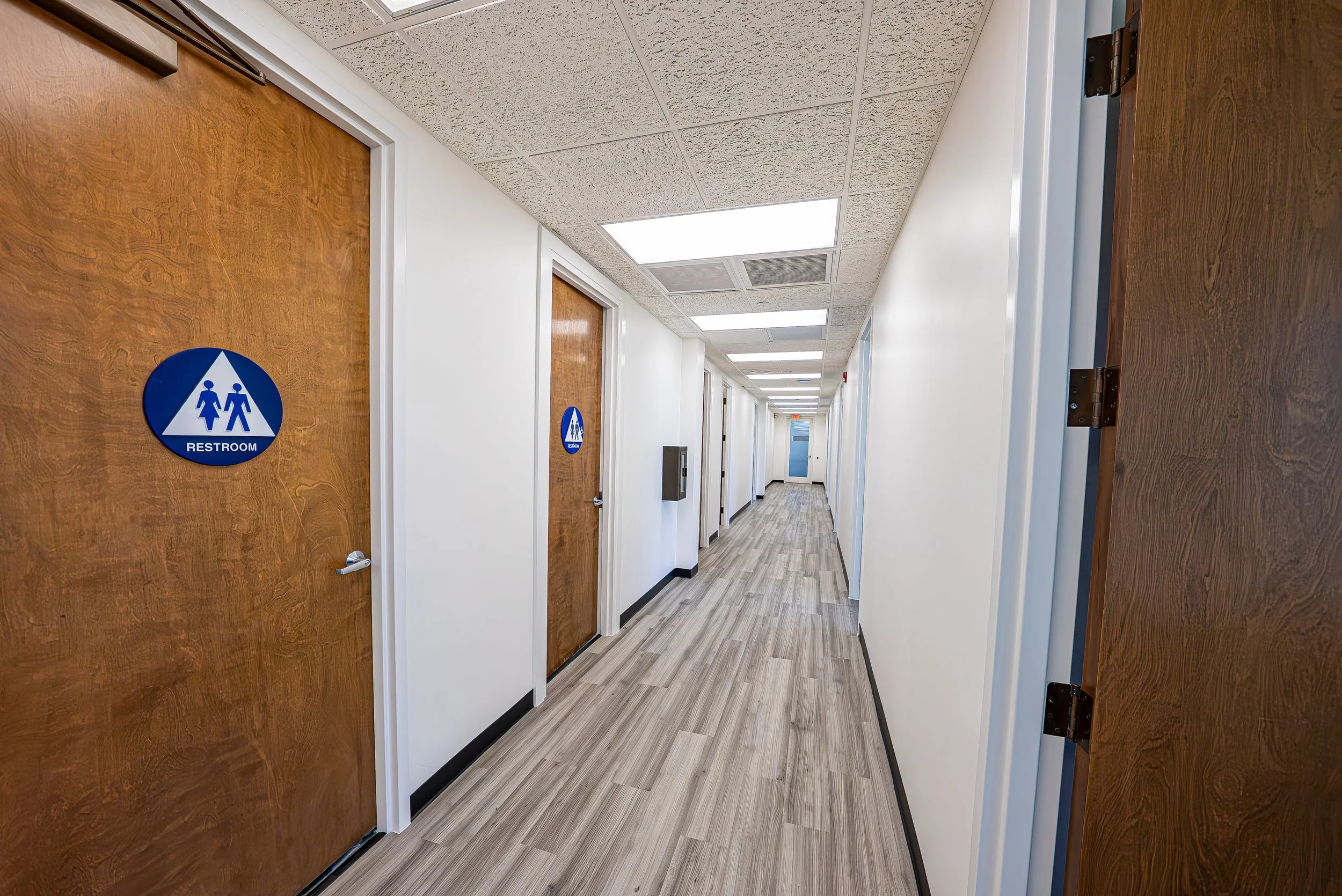 Hallway of an office or medical clinic with multiple restroom doors, each marked with a blue sign, and ceiling lights illuminating the corridor.
