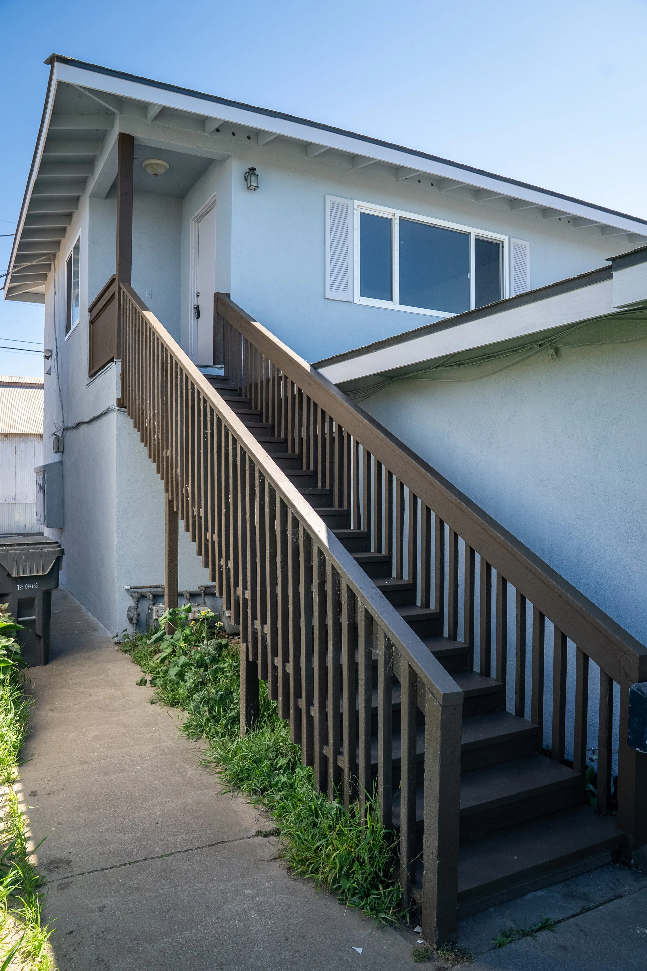 Side view of a light blue two-story house with outdoor stairs leading to the front door, black railing, and a large window with white shutters.