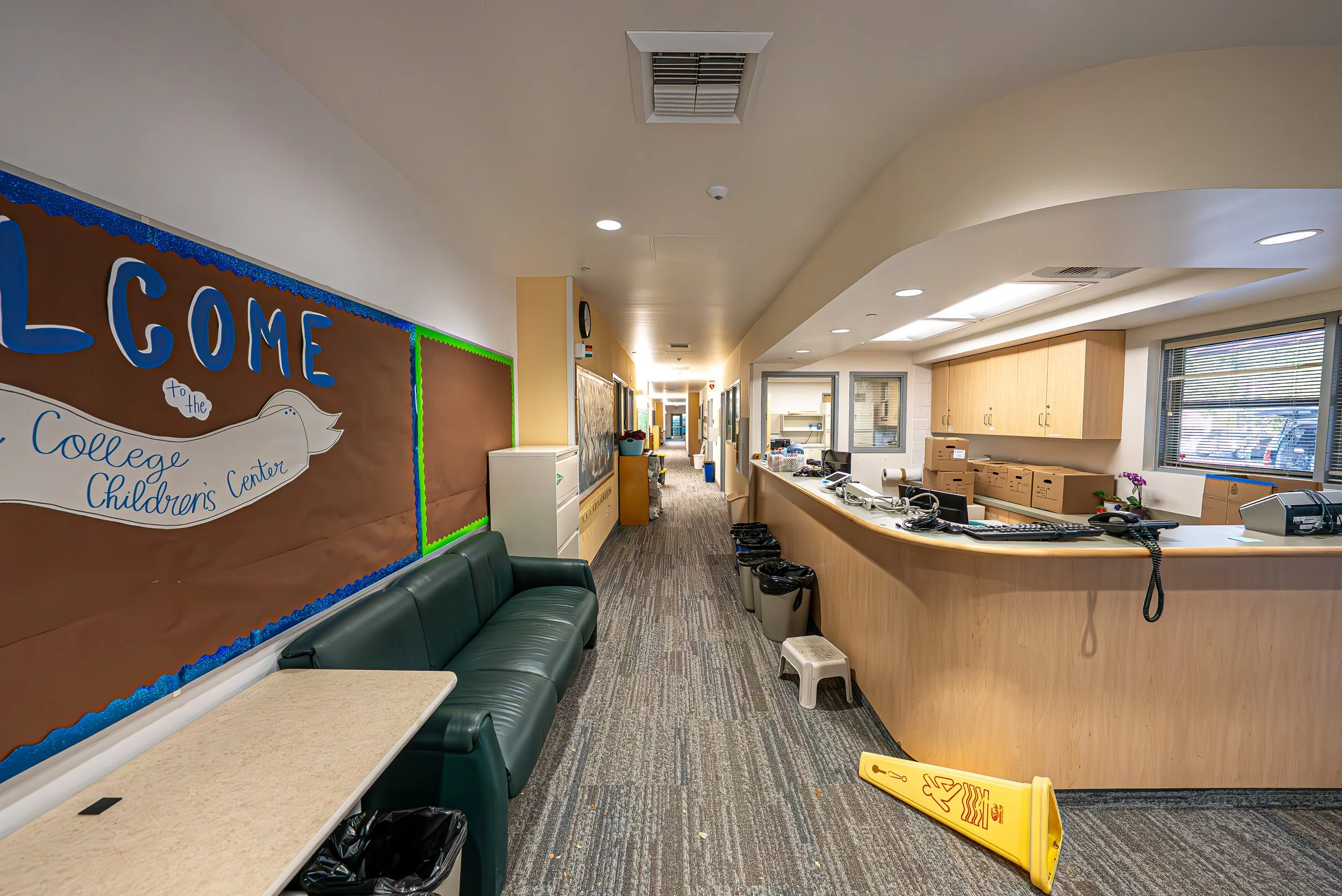 Interior view of a school or healthcare office with a waiting area, colorful bulletin board, reception desk with office supplies, and cleaning supplies on the floor, including a yellow wet floor sign and trash bins.