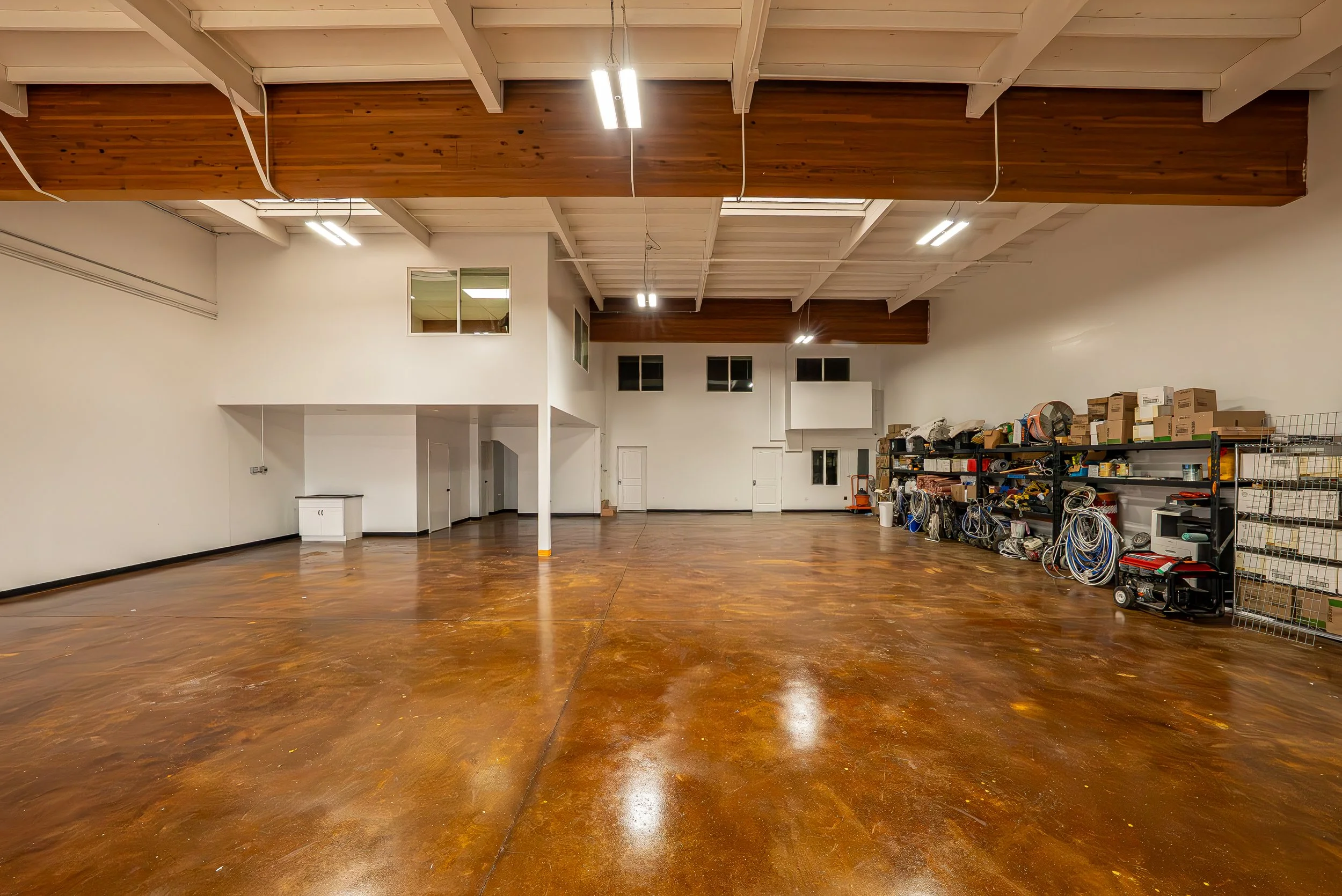 An empty large indoor space with a polished wooden floor, white walls, high ceiling with exposed beams, and bright overhead lighting. Storage shelves with boxes, tools, and equipment are on the right side of the room.