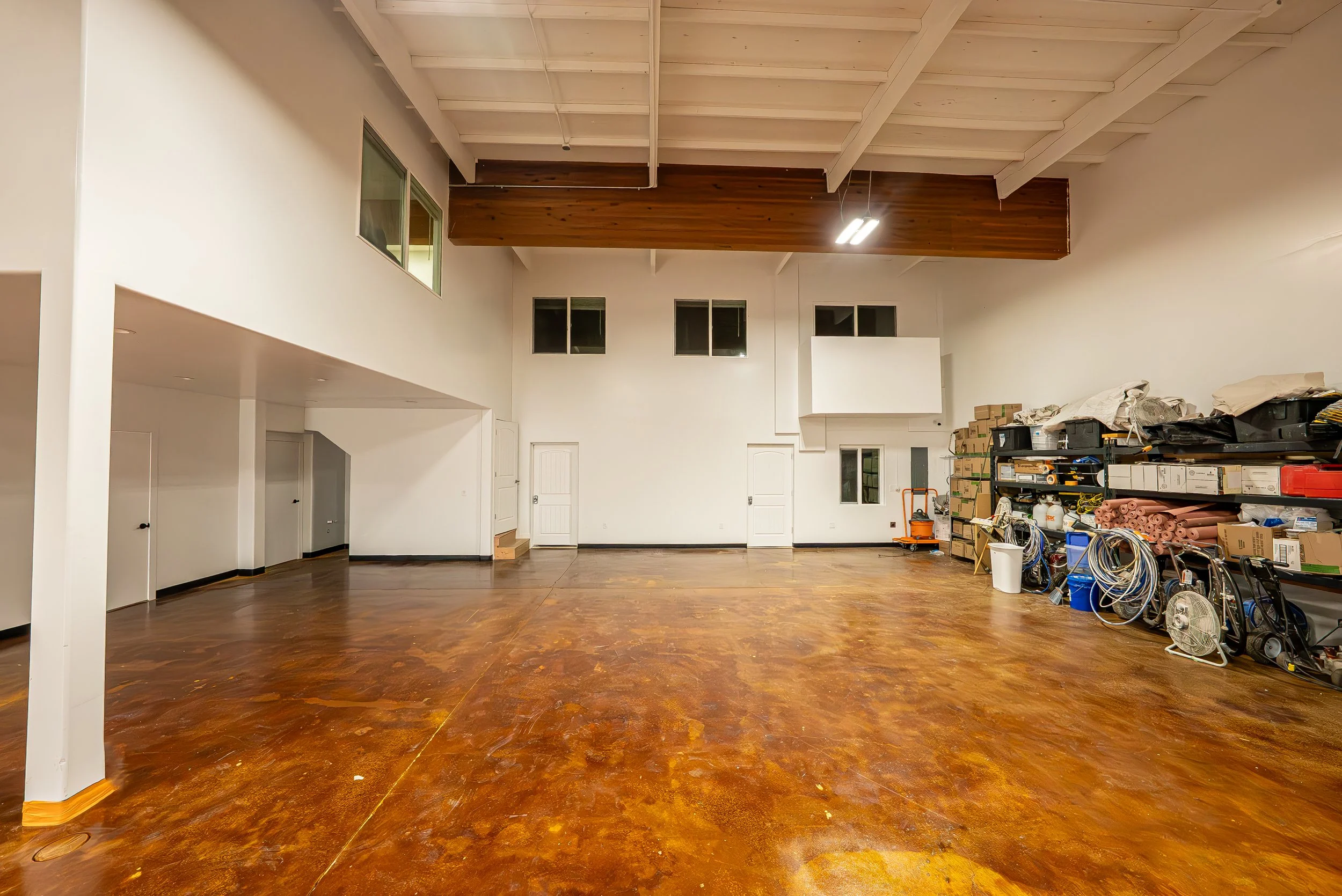 Spacious interior room with polished wooden floor, high white ceiling with exposed beams, white walls with small upper windows, and cluttered storage area with boxes, tools, and equipment on shelving.