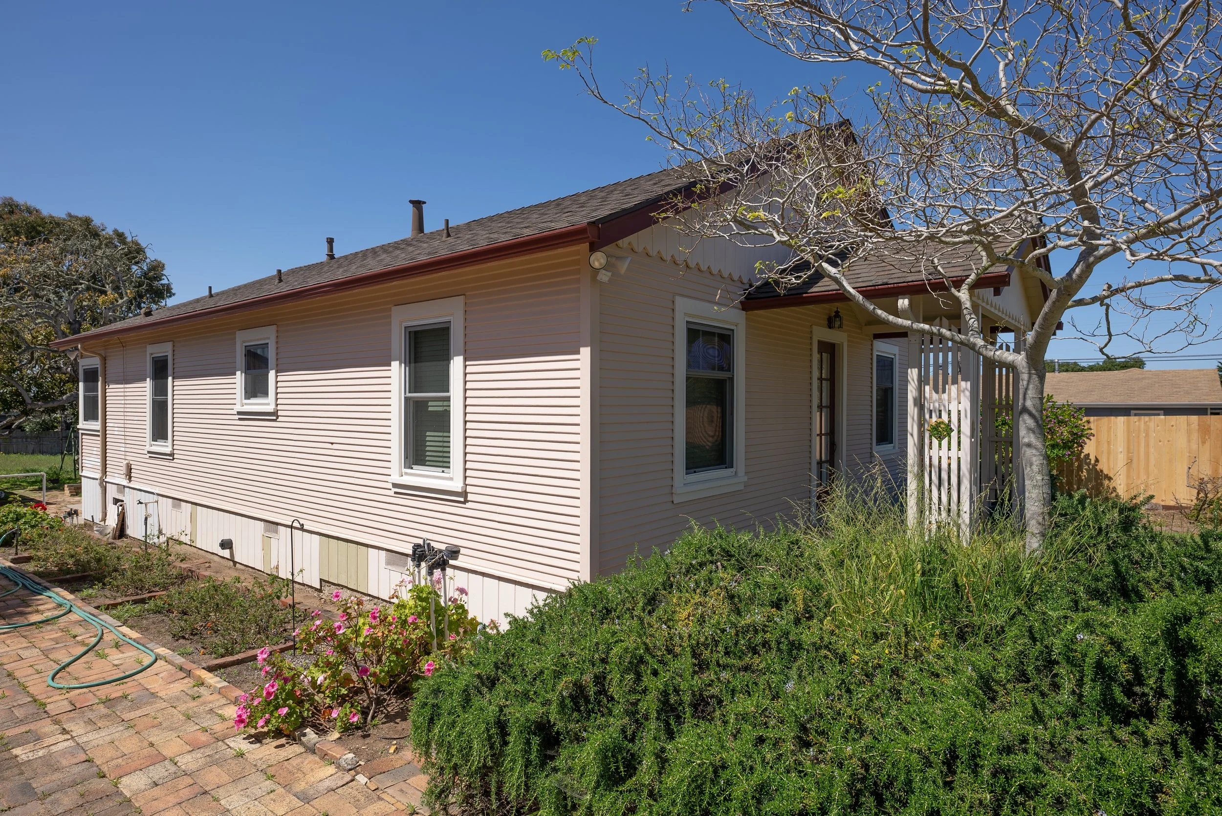 A beige house with white windows and a porch, surrounded by a garden with flowering plants, a brick pathway, and a leafless tree with some green leaves against a clear blue sky.
