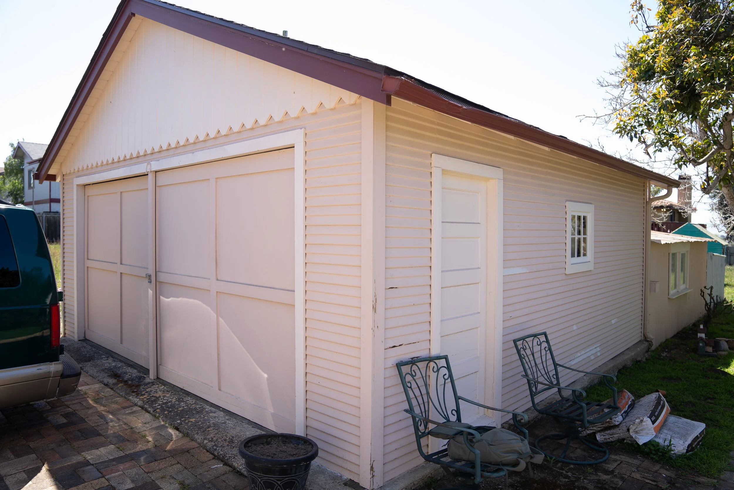 A beige garage with a closed white garage door, a small window on the right side, and a sloped roof. There are two metal chairs and a black pot with a plant in front of the garage, and a pickup truck parked nearby. The yard has brick paving and some 