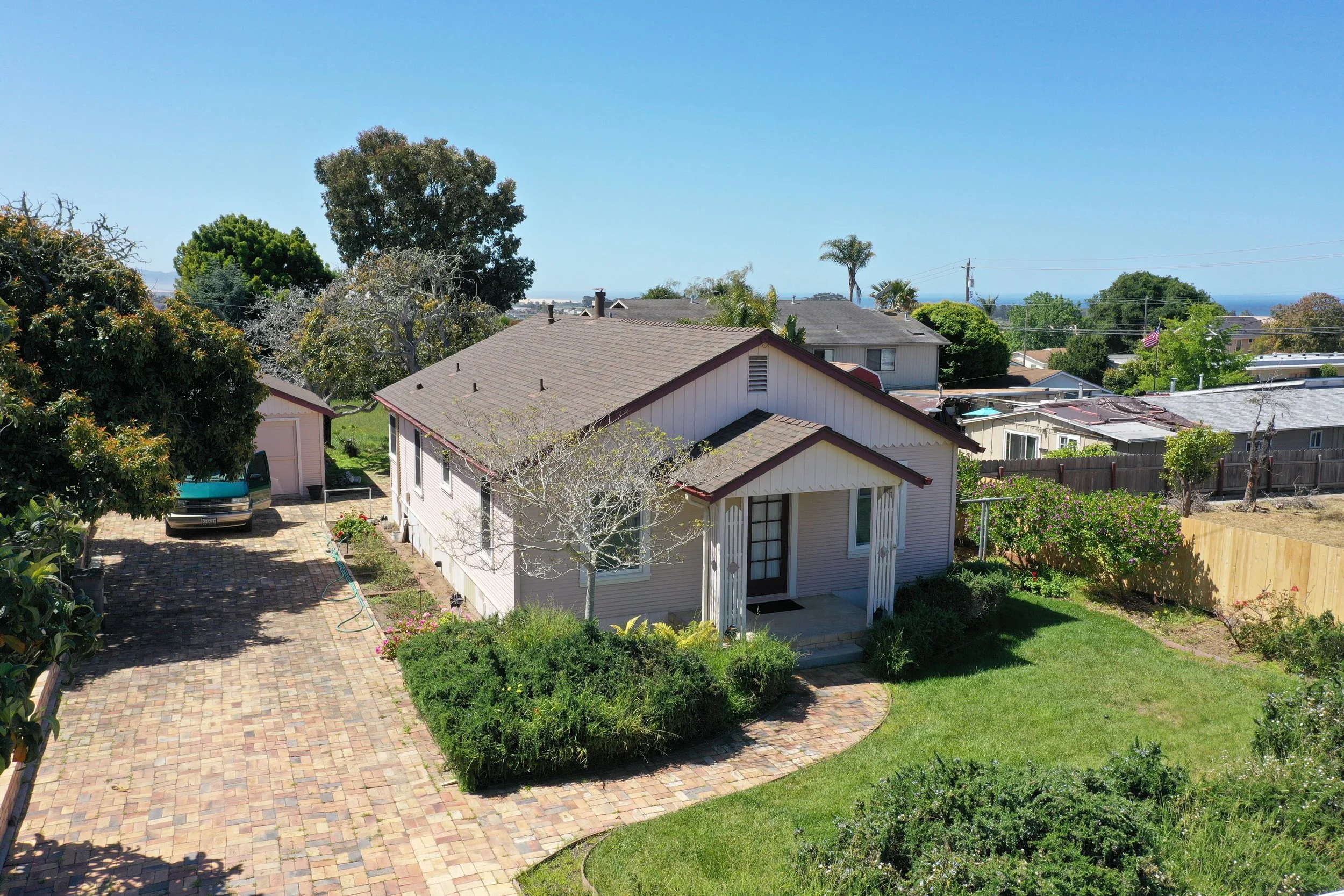 Residential backyard with paved brick patio, lush green grass, various bushes and trees, and houses in the background under a clear blue sky.