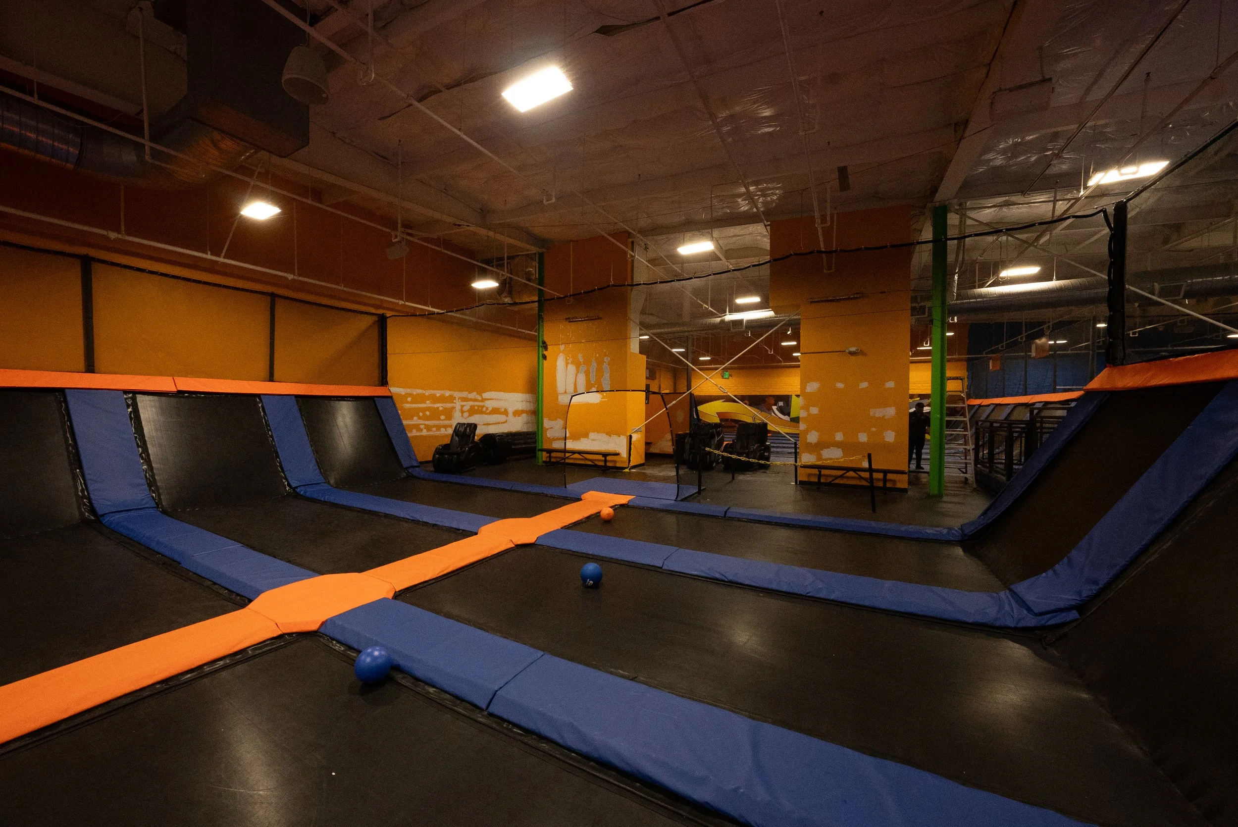 Indoor trampoline park with multiple trampolines, padded walls, and a basketball hoop. There are blue and orange safety pads on the floor, and two balls are visible on the ground.