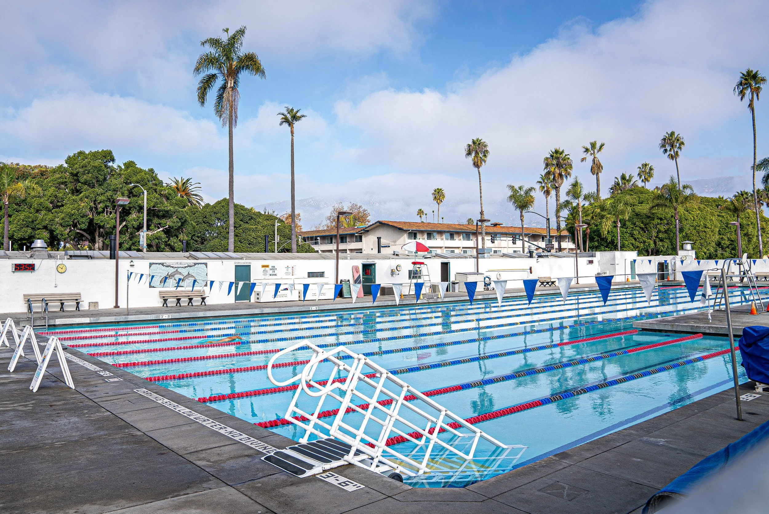 Empty outdoor swimming pool with lane dividers and poolside benches, surrounded by trees and buildings, under a partly cloudy sky.