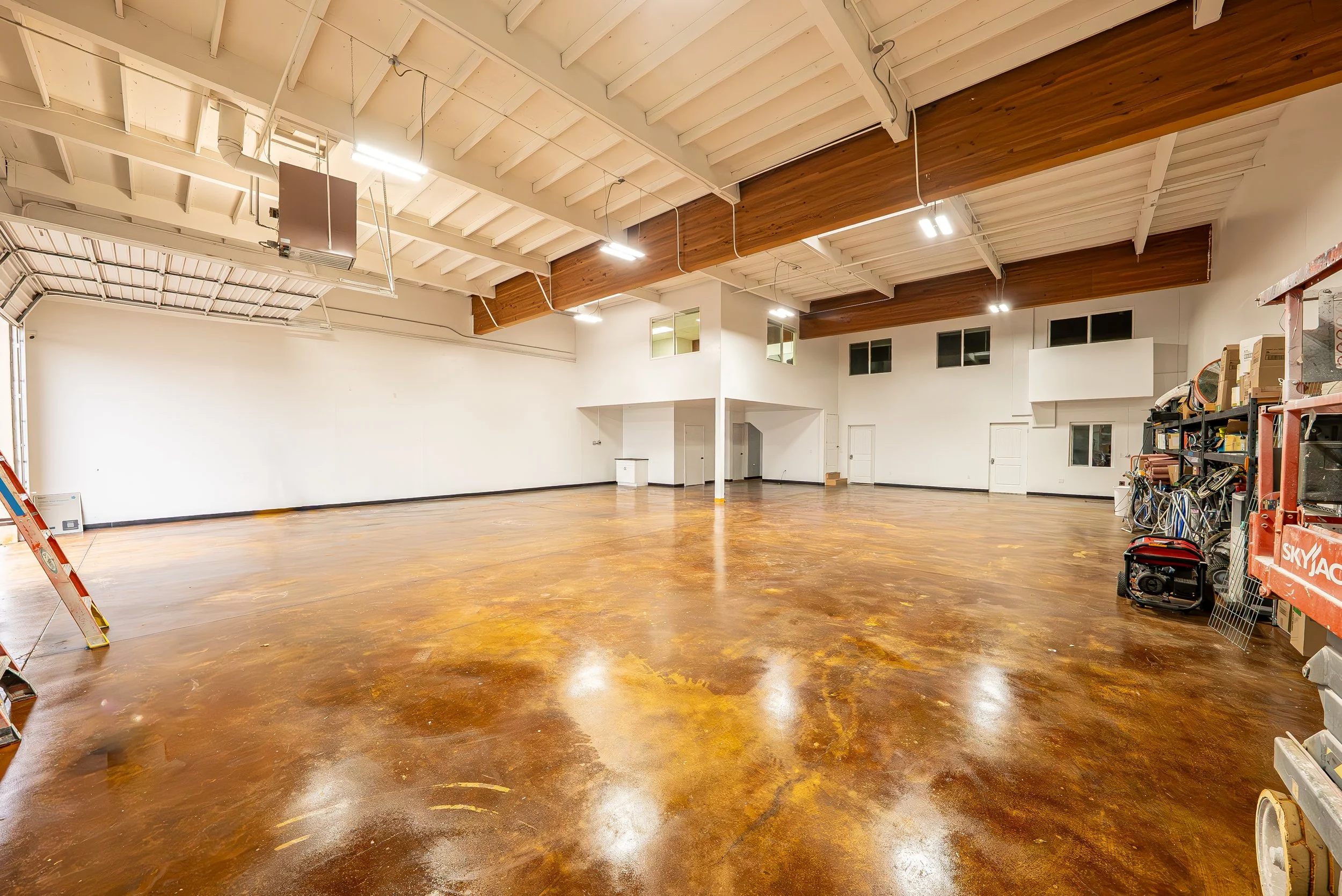 Empty workshop or studio with polished wooden floor, white walls, high ceiling with exposed wooden beams, and shelves with tools and equipment on the right side.