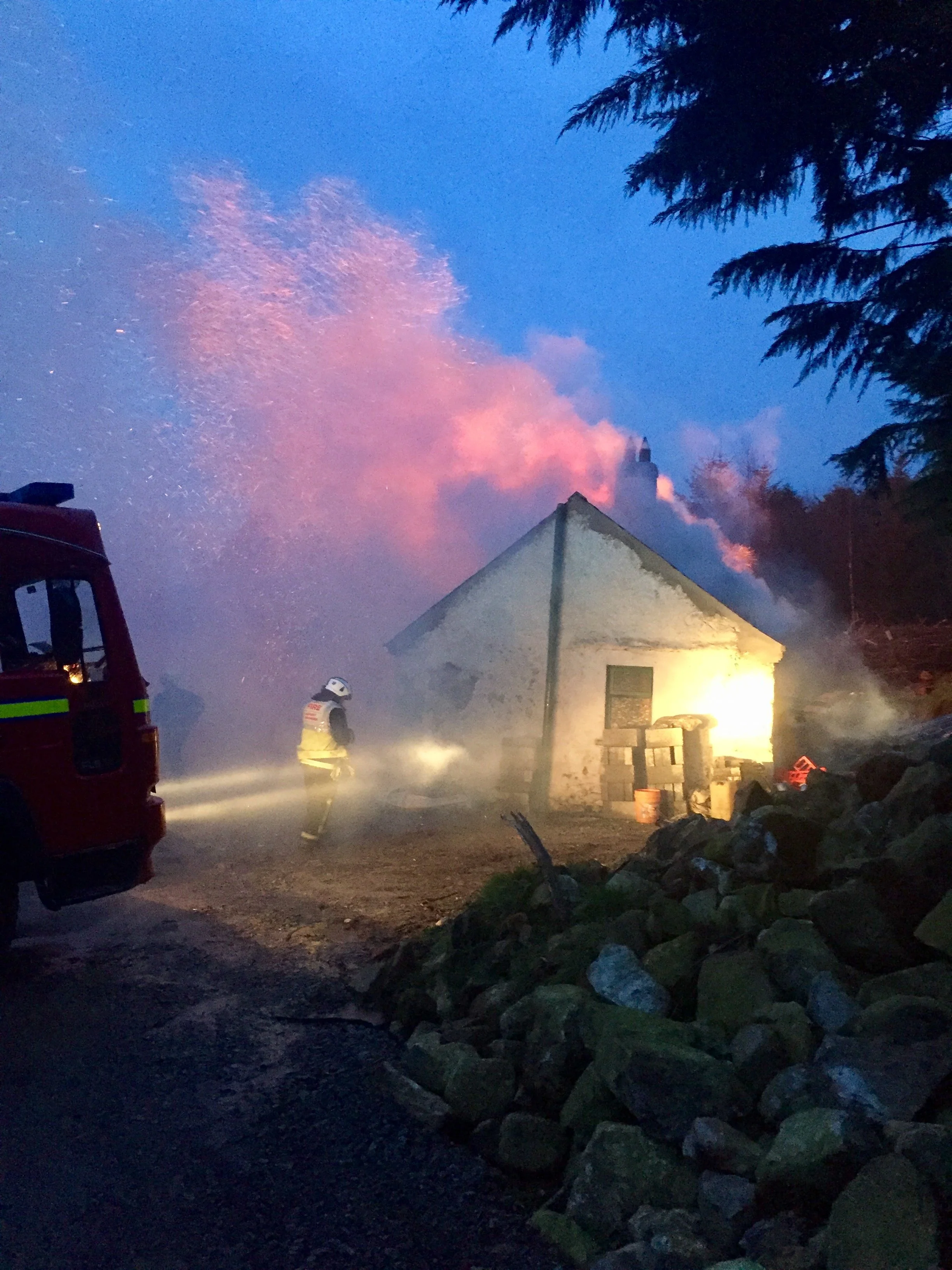 A farmhouse at dusk with smoke near the chimney, warm light from inside, and a fire truck nearby.