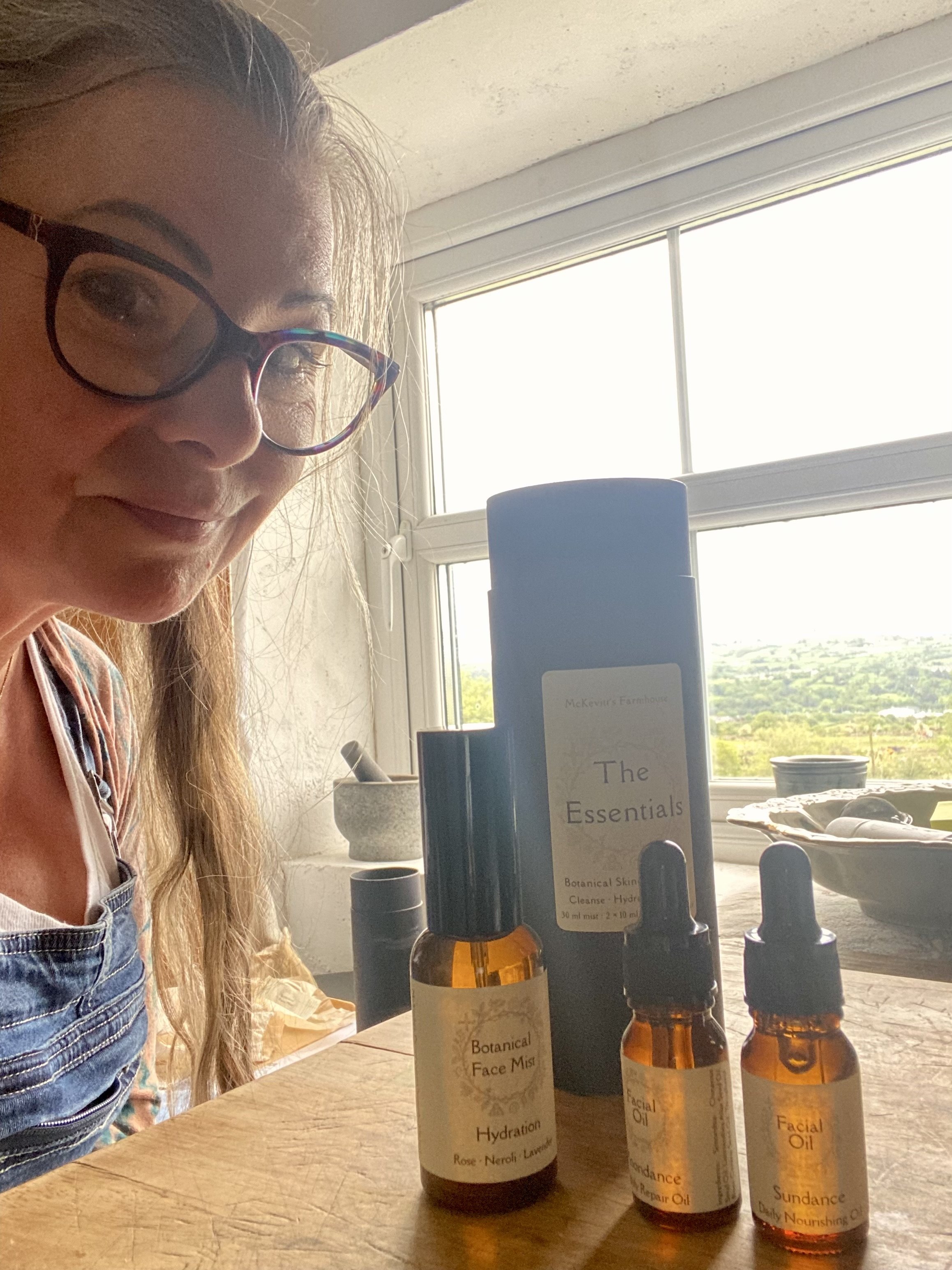 A woman with glasses sitting at a wooden table with several skincare product bottles in front of her, near a window with a scenic view outside.