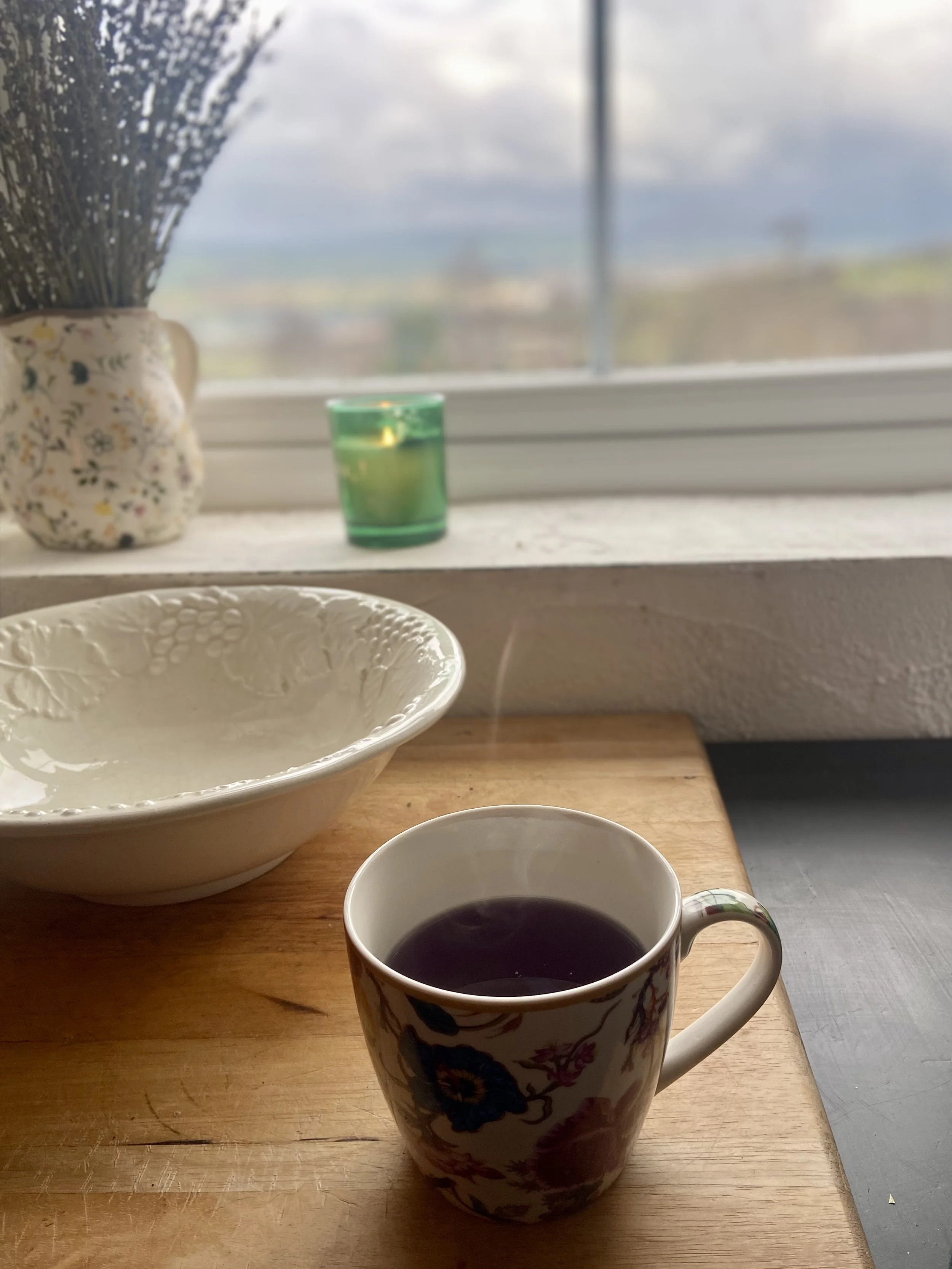 A cup of tea on a wooden table, with a floral-patterned mug, a white bowl, and a ceramic vase with dried flowers in front of a window showing an outdoor view.
