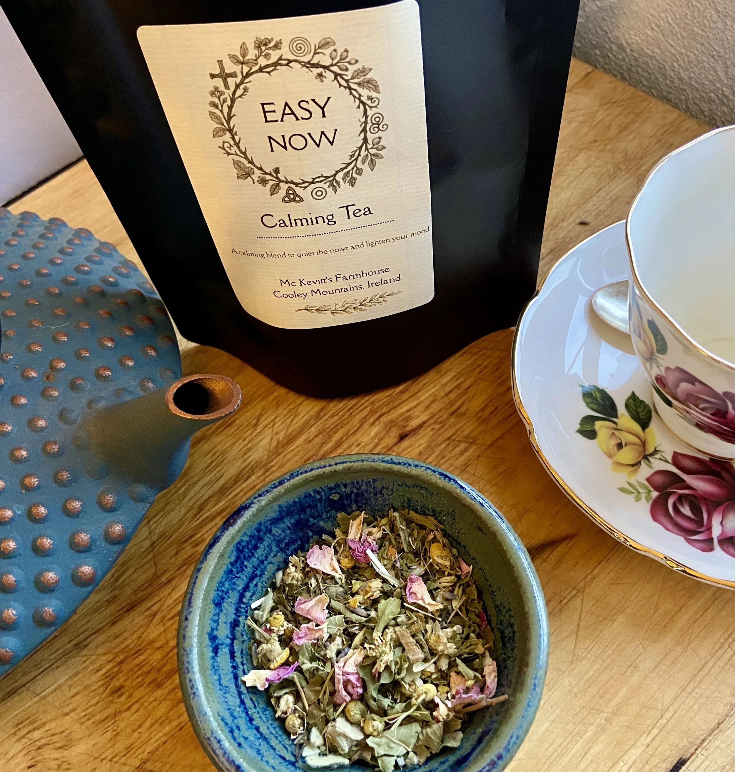 Close-up of a ceramic bowl filled with dried herbal tea herbs on a wooden surface. Part of a decorative teacup with floral design is visible on the right. In the background, a package labeled "Easy Now" Calming Tea from McKevitt's Farmhouse in Cooley Mountains, Ireland, is visible.