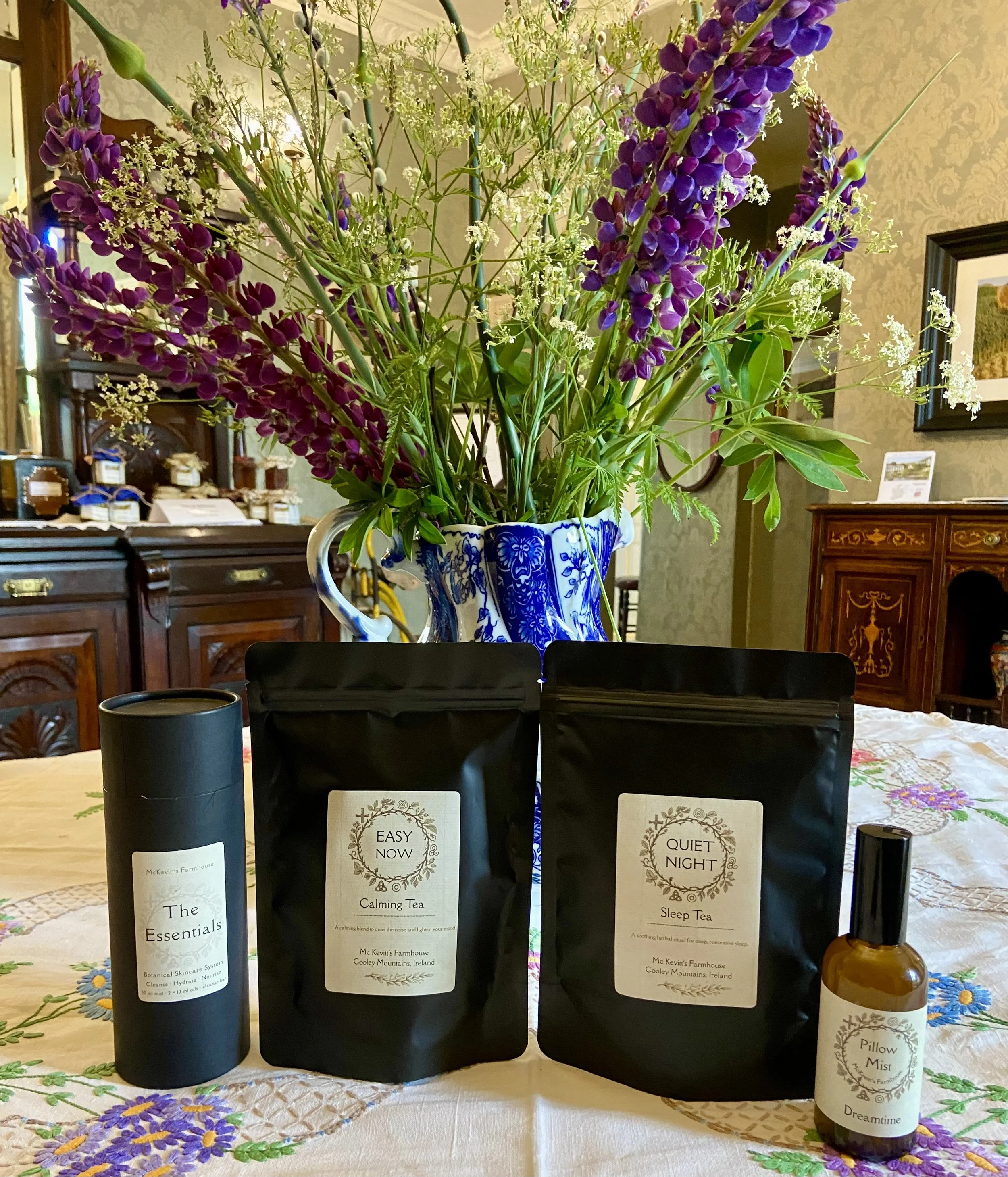 A bouquet of purple and white flowers in a decorative blue and white vase on a table. In front of the bouquet, there are three bottles of herbal tea with labels reading "The Essentials", "Calming Tea", and "Sleep Tea." The tablecloth has a floral embroidered pattern.