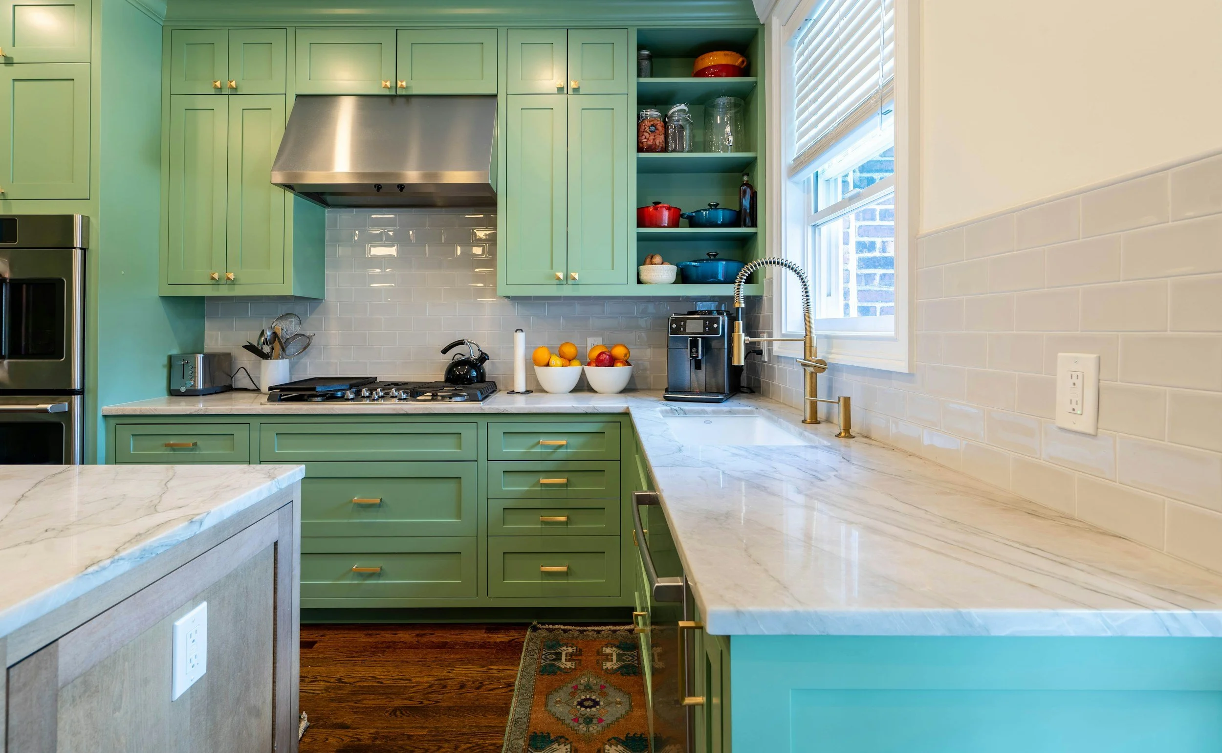 Kitchen with green cabinets, white subway tile backsplash, marble countertops, and a window. Contains a stove, coffee maker, bowls of fruit, and open shelves with pots and jars.