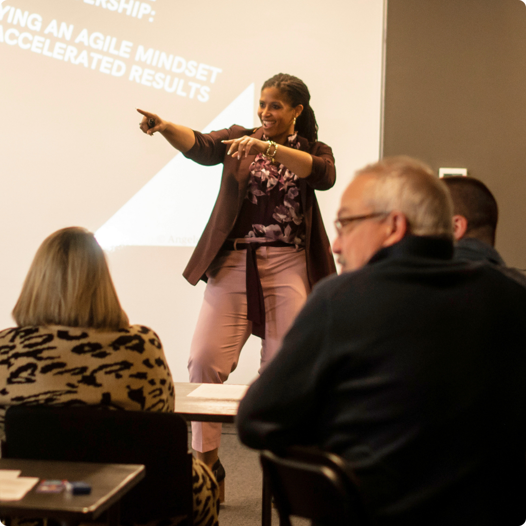 A woman giving a presentation in a conference room, pointing towards a projected slide with text on it, while three people seated at a table listen.