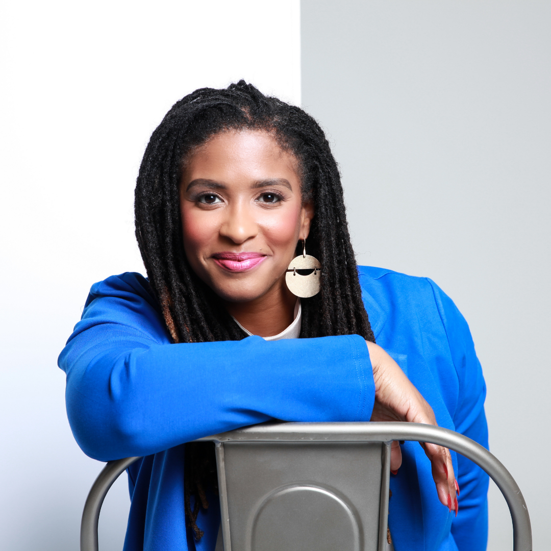 A woman with dark dreadlocks, wearing a royal blue blazer and large gold earrings, leaning on a metal chair with a light gray wall in the background.
