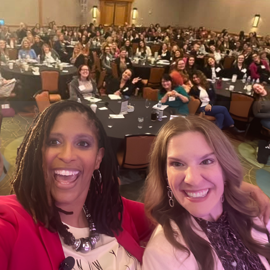 Two women taking a selfie in front of a large group of people seated at banquet tables in a conference room.