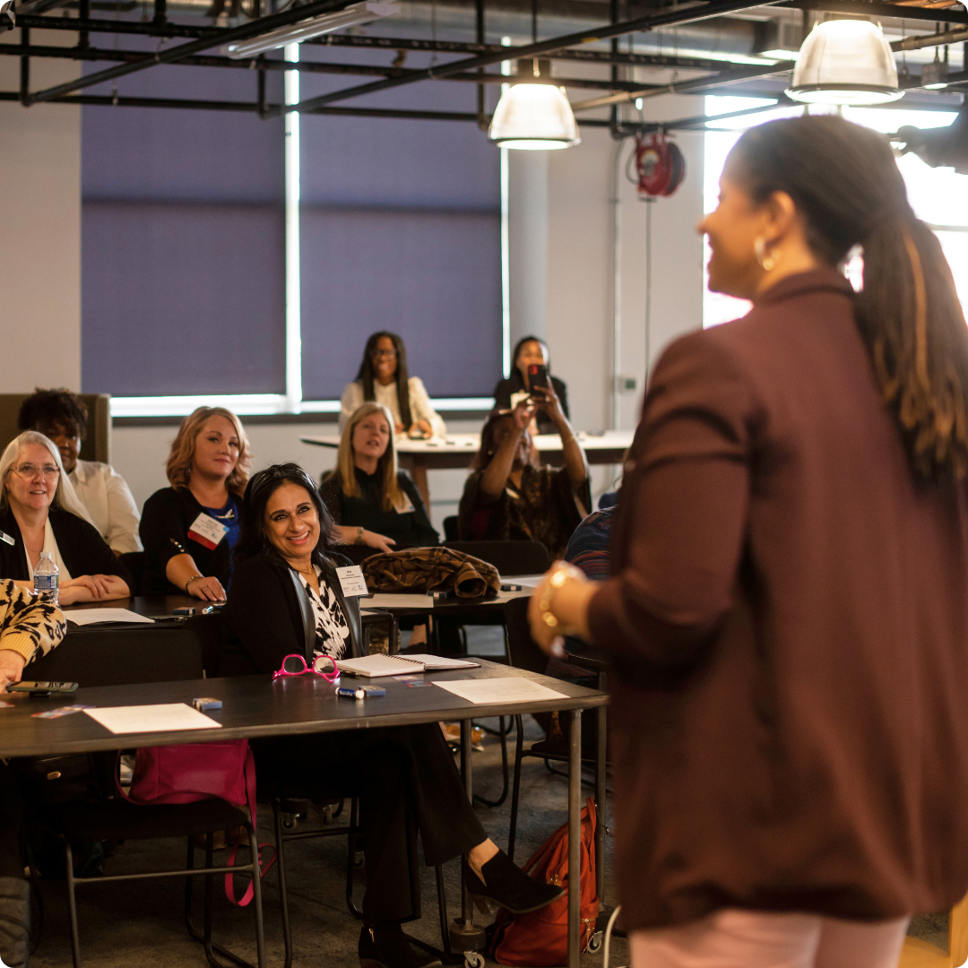 A woman is giving a presentation to a group of people seated at tables in a room with industrial lighting and large windows covered with blinds.