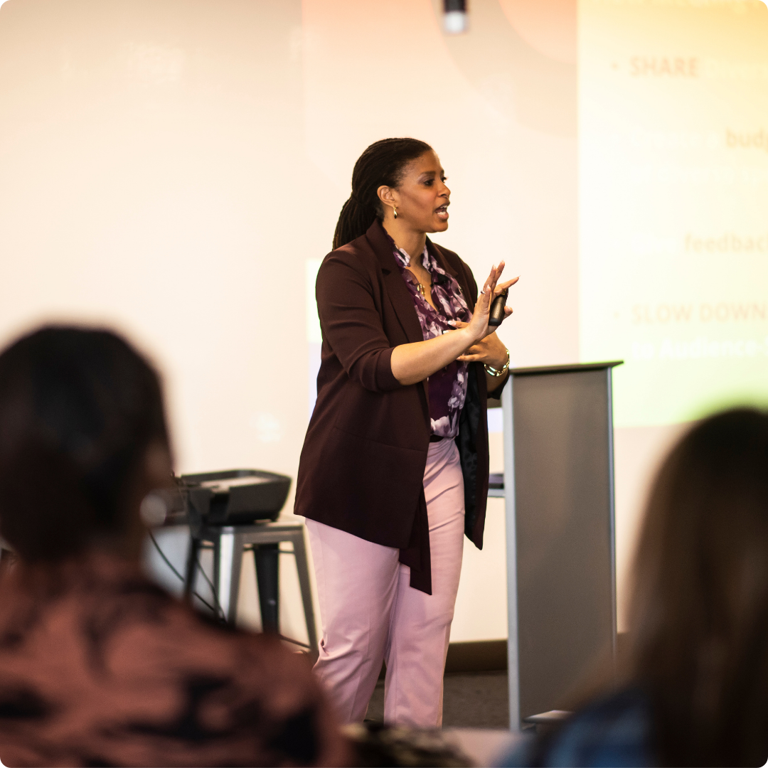 A woman giving a presentation in a conference room, standing beside a podium, with a projected slide in the background.