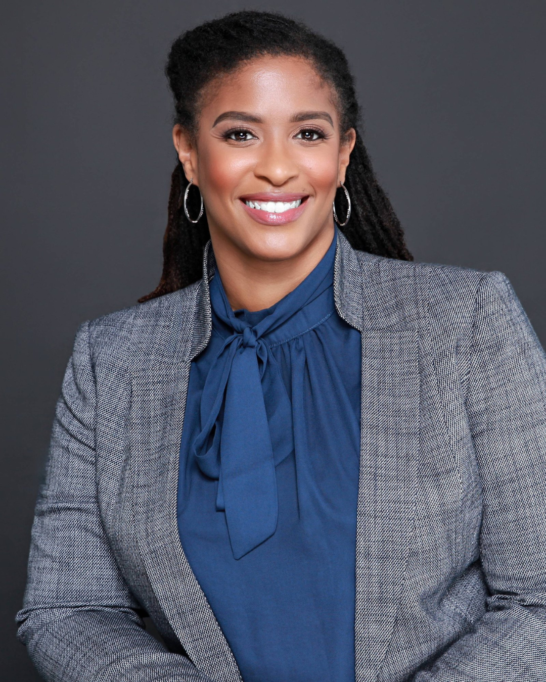 Professional portrait of a smiling African American woman with long dreadlocks, wearing a blue- gray blazer and blue blouse with a bow, against a dark gray background.