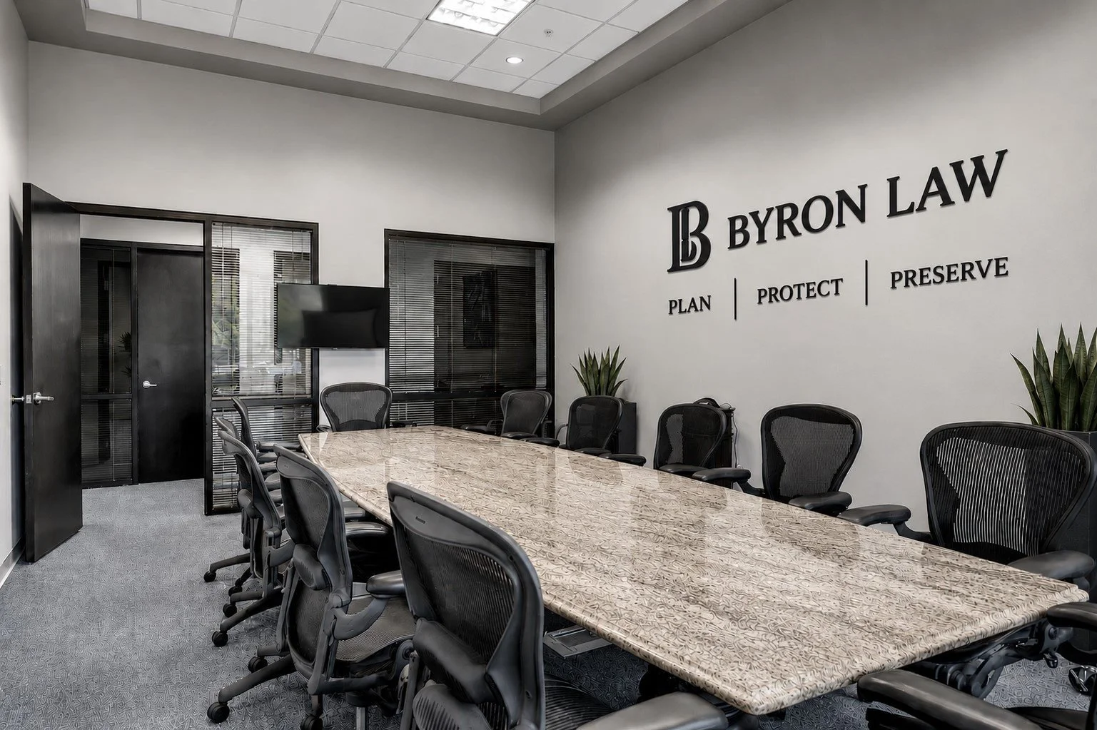 A modern conference room with a large marble table, black swivel chairs, a wall-mounted TV, and a wall with the Byron Law logo and the words 'Plan', 'Protect', and 'Preserve'.