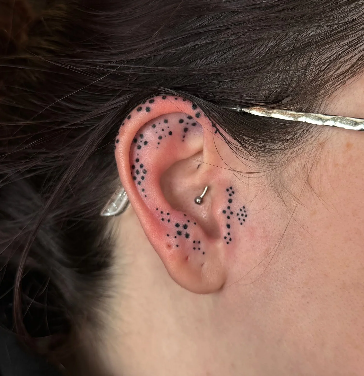 Close-up of a person's left ear with multiple black dots tattooed around the outer ear and the earlobe. Custom Ear Curation.