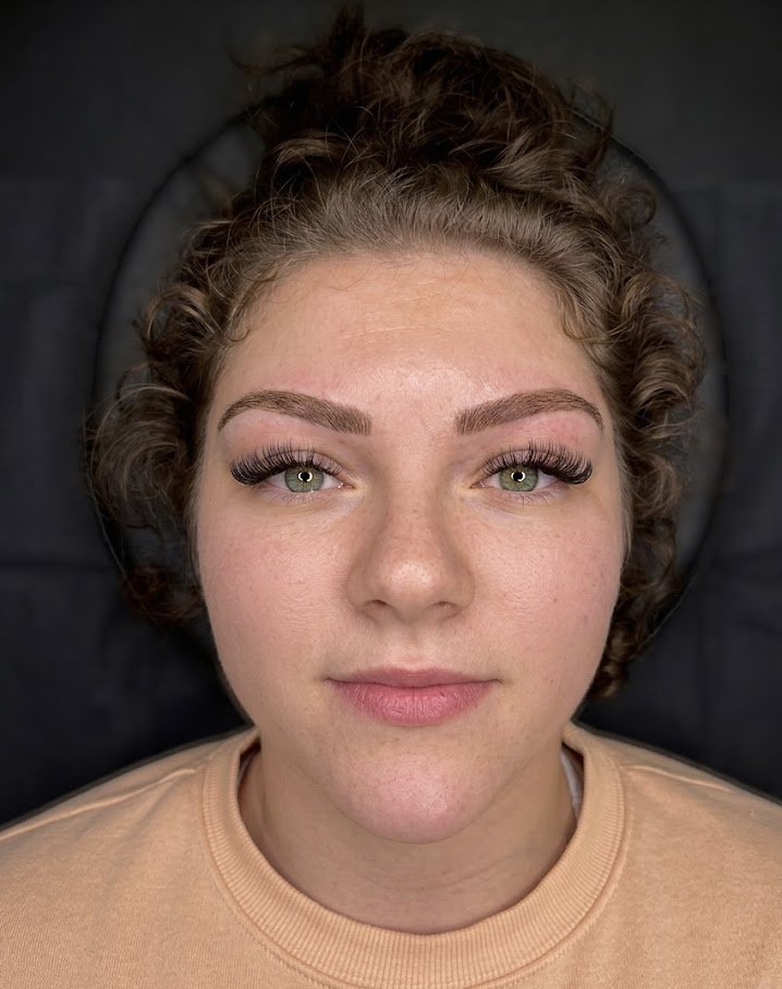 Close-up of a young woman with curly hair and light makeup, lying down with her head against a black background. Hybrid brow.