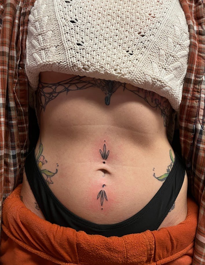 Close-up of a person's midsection showing tattoos on the stomach and hips, with a crocheted sweater partially covering the chest and a flannel shirt on the sides.