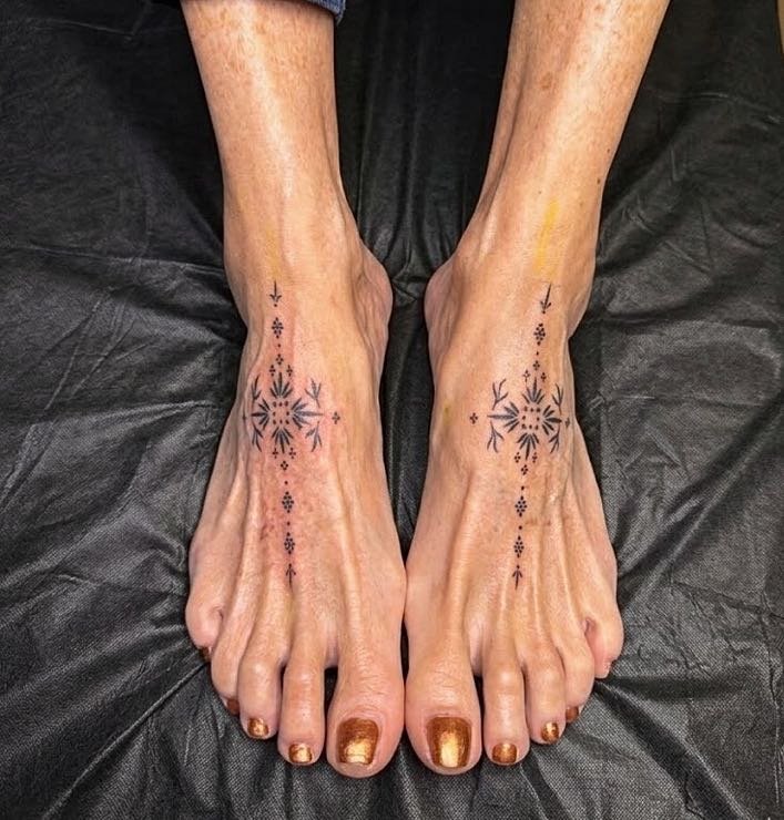 Close-up of person's feet with tattoos on the top, resting on a black surface. The tattoos are symmetrical, black, and intricate in design. Custom Feet Curation.