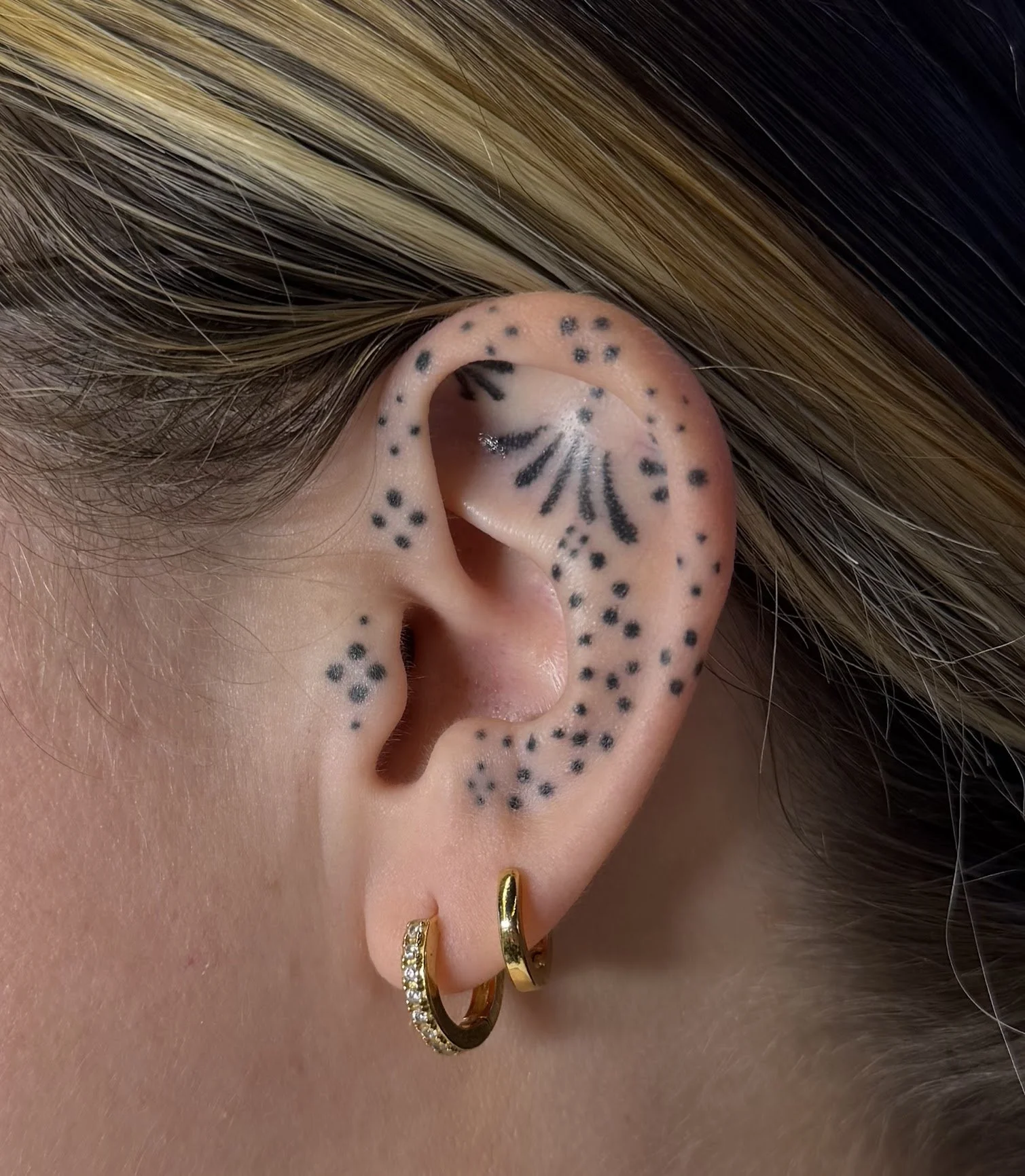 Close-up of a person's ear decorated with multiple black dots and lines of tattoo ink, along with gold hoop earrings and light brown hair. Custom Ear Curation.