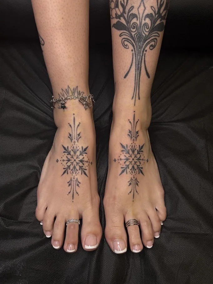 Tattooed feet with black ink symmetrical designs, floral and abstract patterns, on a black surface. Custom Feet Curation.