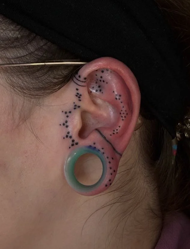 Close-up of a person's ear with a focus on multiple black dots marked around the ear, a large turquoise and green gauge earring in the earlobe. Custom Ear Curation.