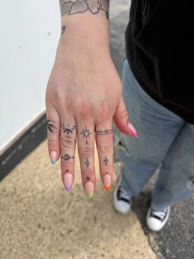 A person's hand with colorful, rainbow-gradient painted nails, displaying multiple small tattoos on the fingers, including stars, a moon, and ornamental designs.