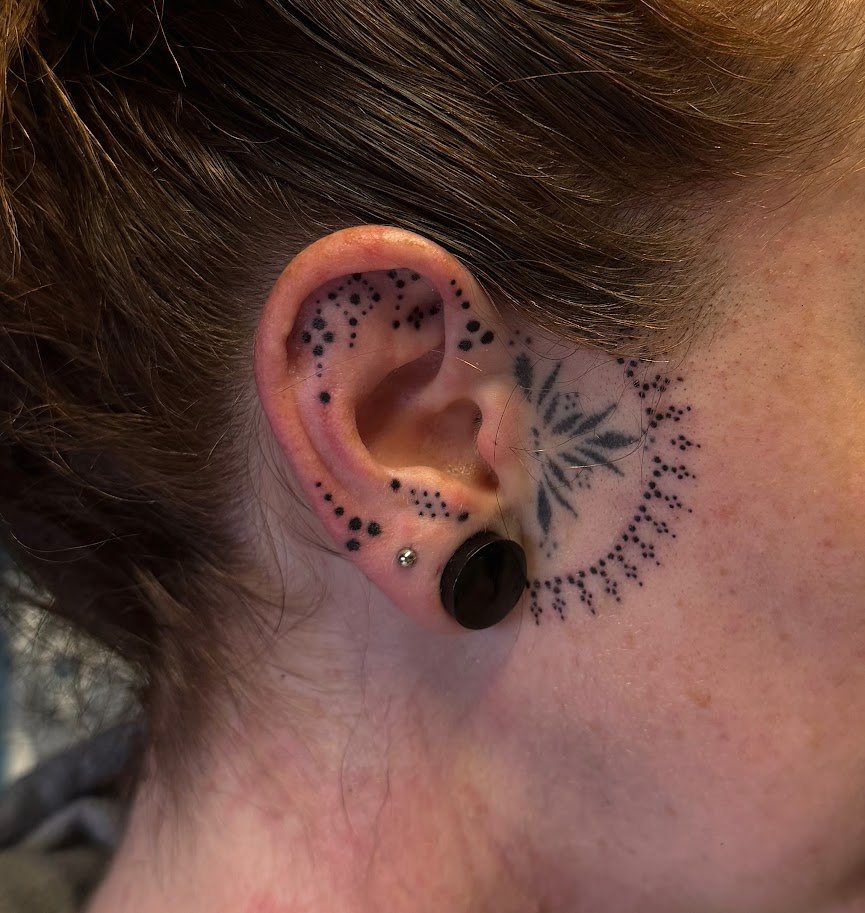 Close-up of a person's ear with multiple piercings and tattoos, including black dots and a black leaf design behind the ear.