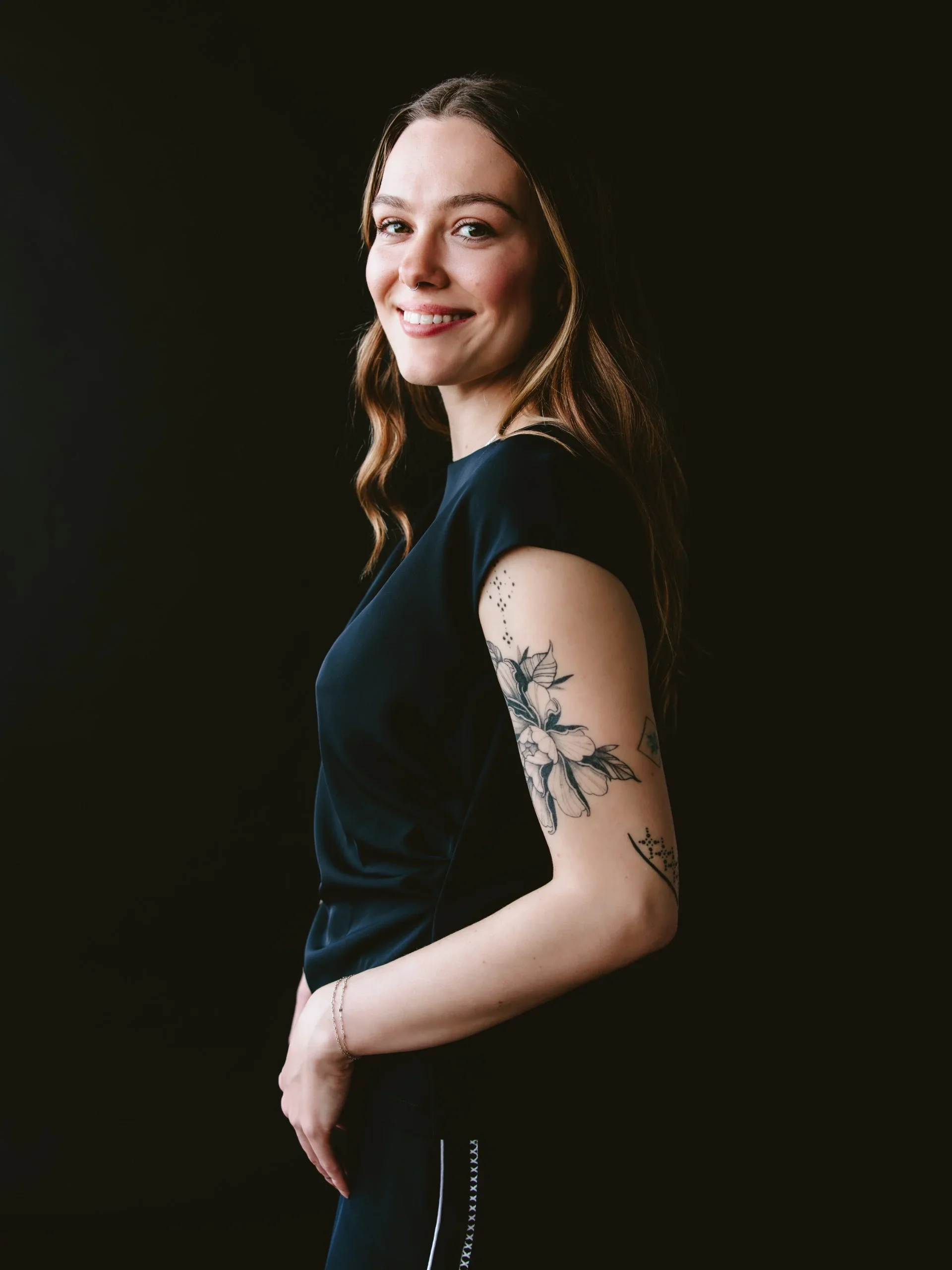 A young woman with long brown hair and a nose ring, smiling and looking at the camera, wearing a black dress with tattoos on her right arm, against a dark background.