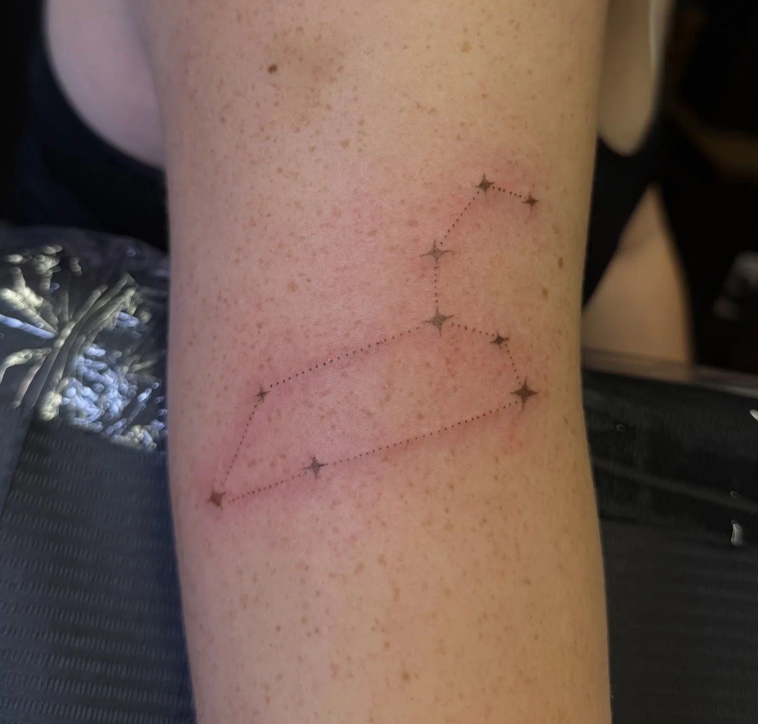 A person's arm with a dotted outline of a constellation tattoo, featuring star symbols connected by dashed lines.