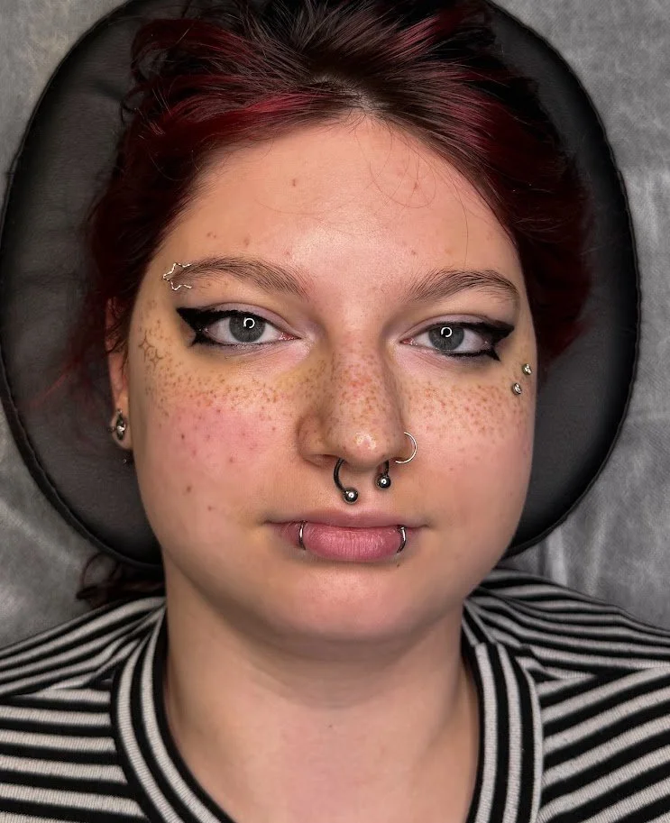 A close-up of a young woman with red hair and numerous facial piercings, lying on a pillow with a striped shirt.