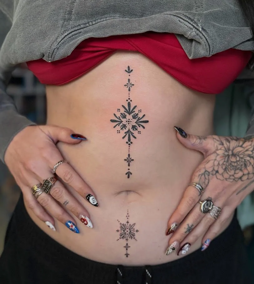 Tattooed midsection of a person with hands on hips, showing a symmetrical floral tattoo design on the stomach and a smaller matching design on the lower abdomen, with colorful nail art and rings on fingers, wearing a red top and a gray hoodie.