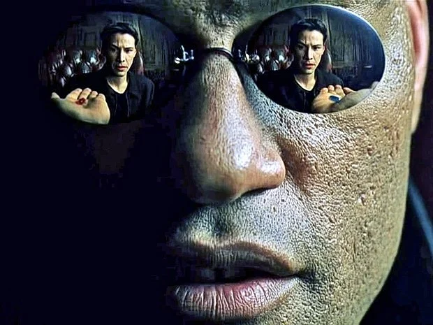 Close-up of a person's face wearing reflective sunglasses, showing the reflection of a woman with red nail polish in the lenses.